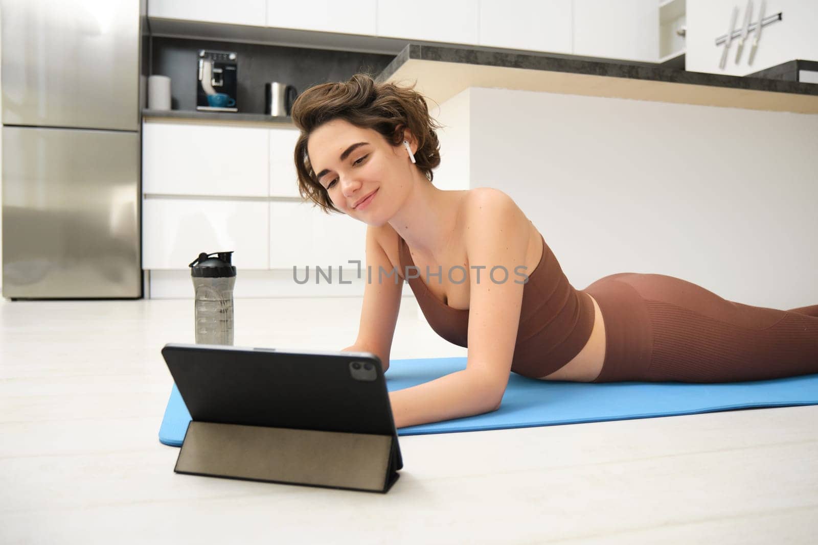 Full body smiling young strong sporty athletic fitness trainer instructor woman wear brown tracksuit use tablet for online training do exercises at home gym indoor. Workout sport concept.