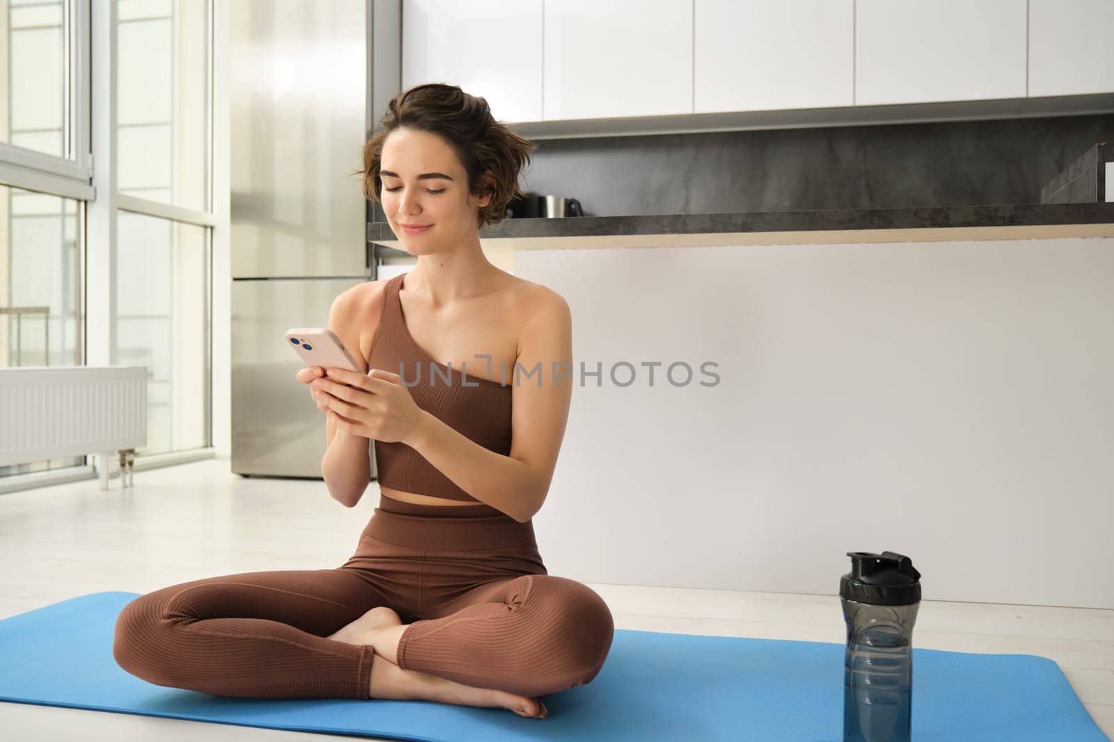 Portrait of girl doing yoga at home, using smartphone app and smiling, tracking workout progress on mobile phone. Sport and wellbeing concept