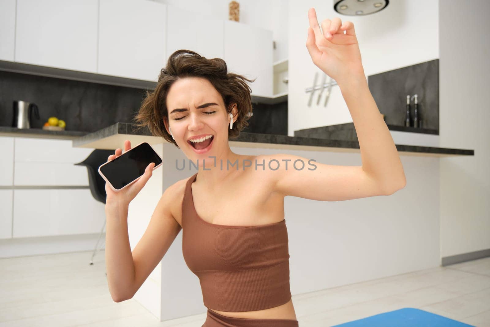 Portrait of brunette girl, does workout at home, sings in smartphone, listens music in wireless headphones while doing exercises, fitness training on kitchen floor, sport and healthy lifestyle.