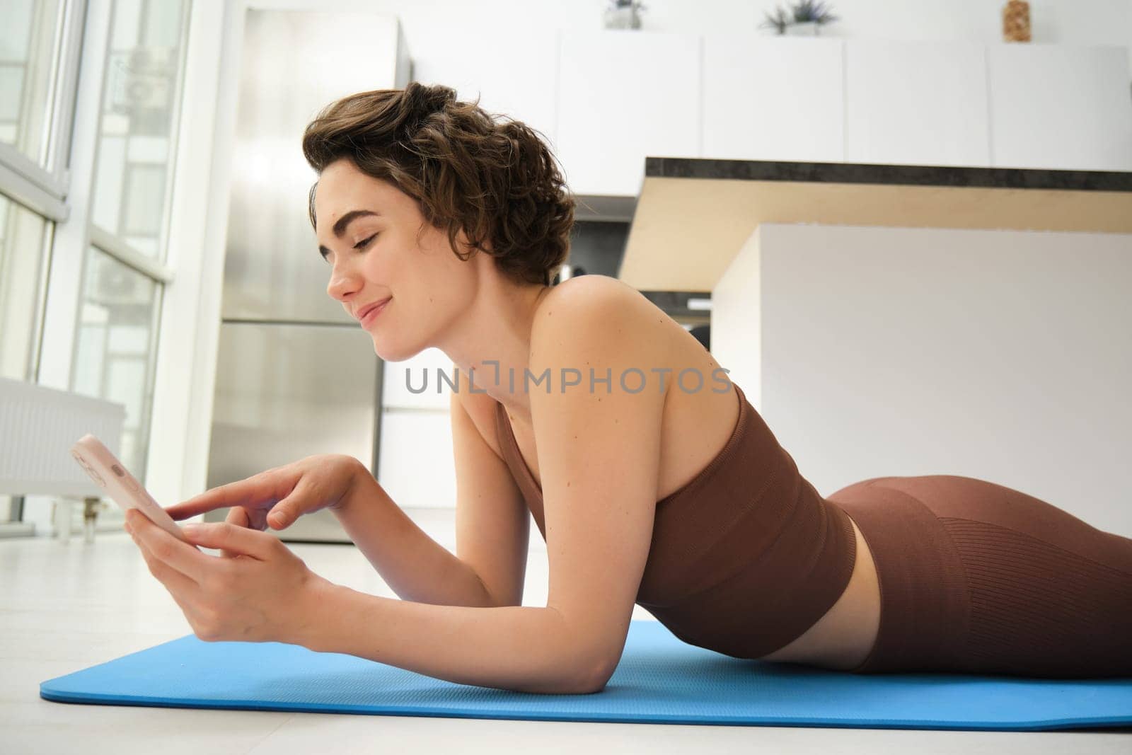 Indoor shot of fit and healthy woman workout at home, lying on yoga mat while using smartphone, watching fitness training at mobile phone, resting after exercises.