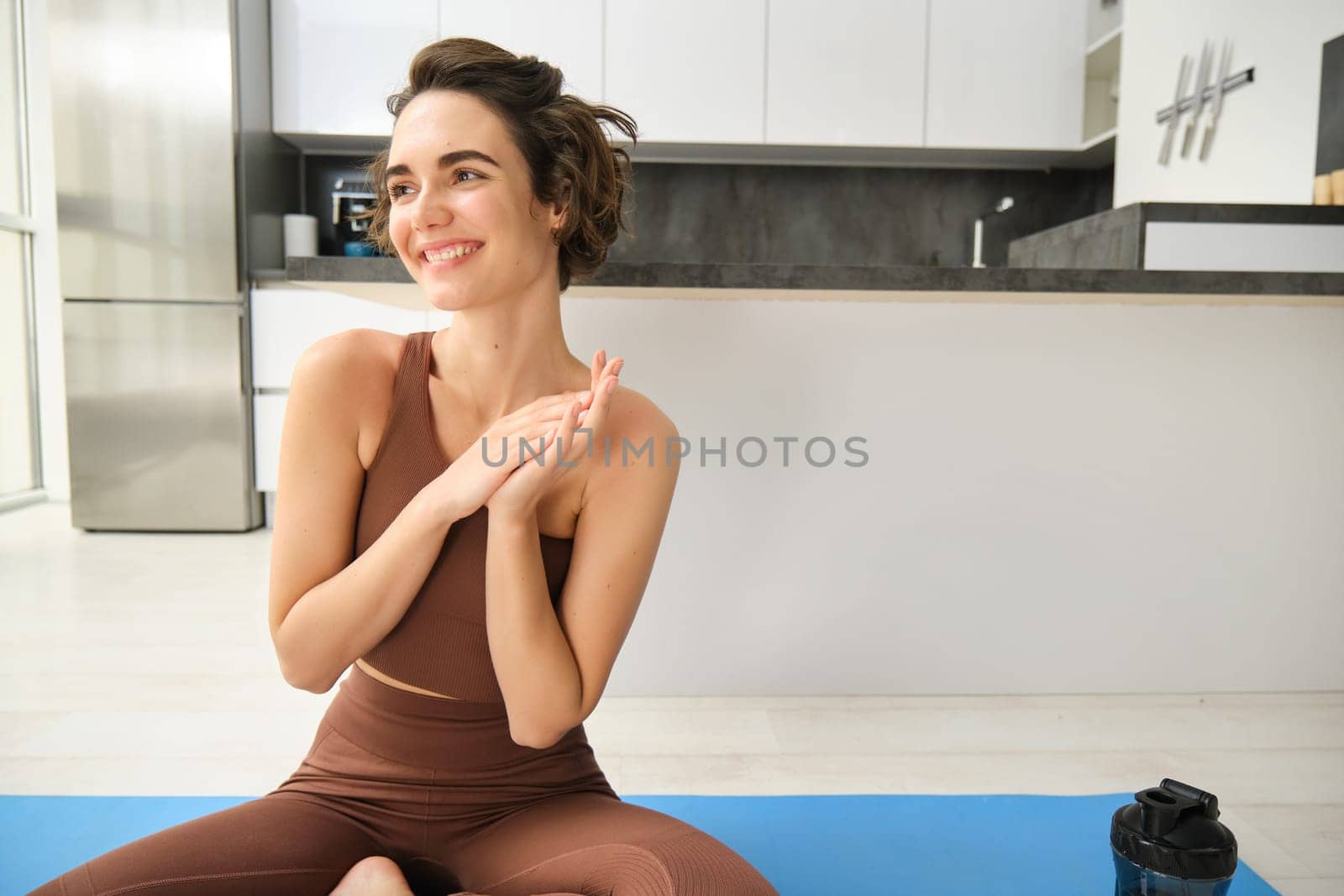 Happy and relaxed fitness girl, sitting on rubber mat doing yoga, meditating at home, doing mindfulness practice indoors. Sport and wellbeing concept