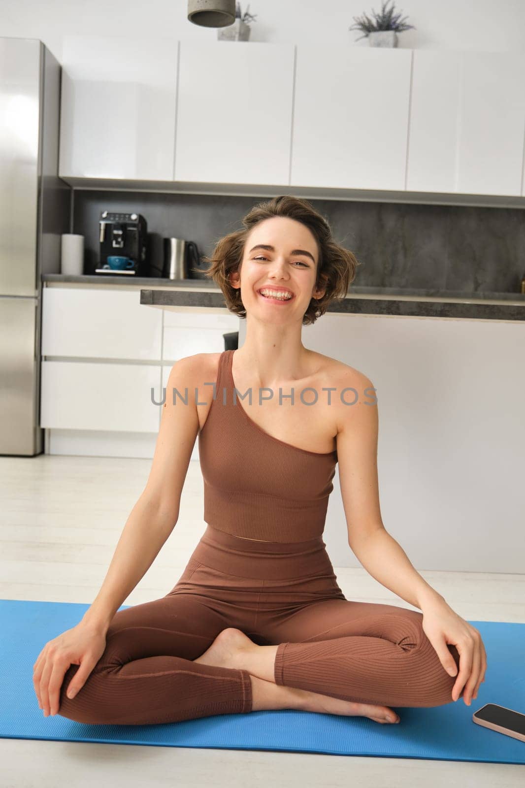 Vertical shot of young woman sitting at home on floor, doing yoga, making lotus pose and meditating, feeling relaxed and happy, smiling by Benzoix