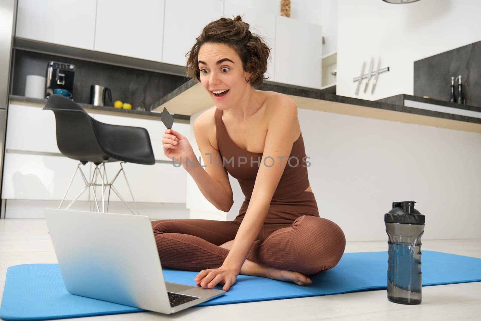 Image of young fitness girl, female athlete at home, workout on kitchen floor, holding credit card with laptop, purchasing online training session, gym application for home workout by Benzoix