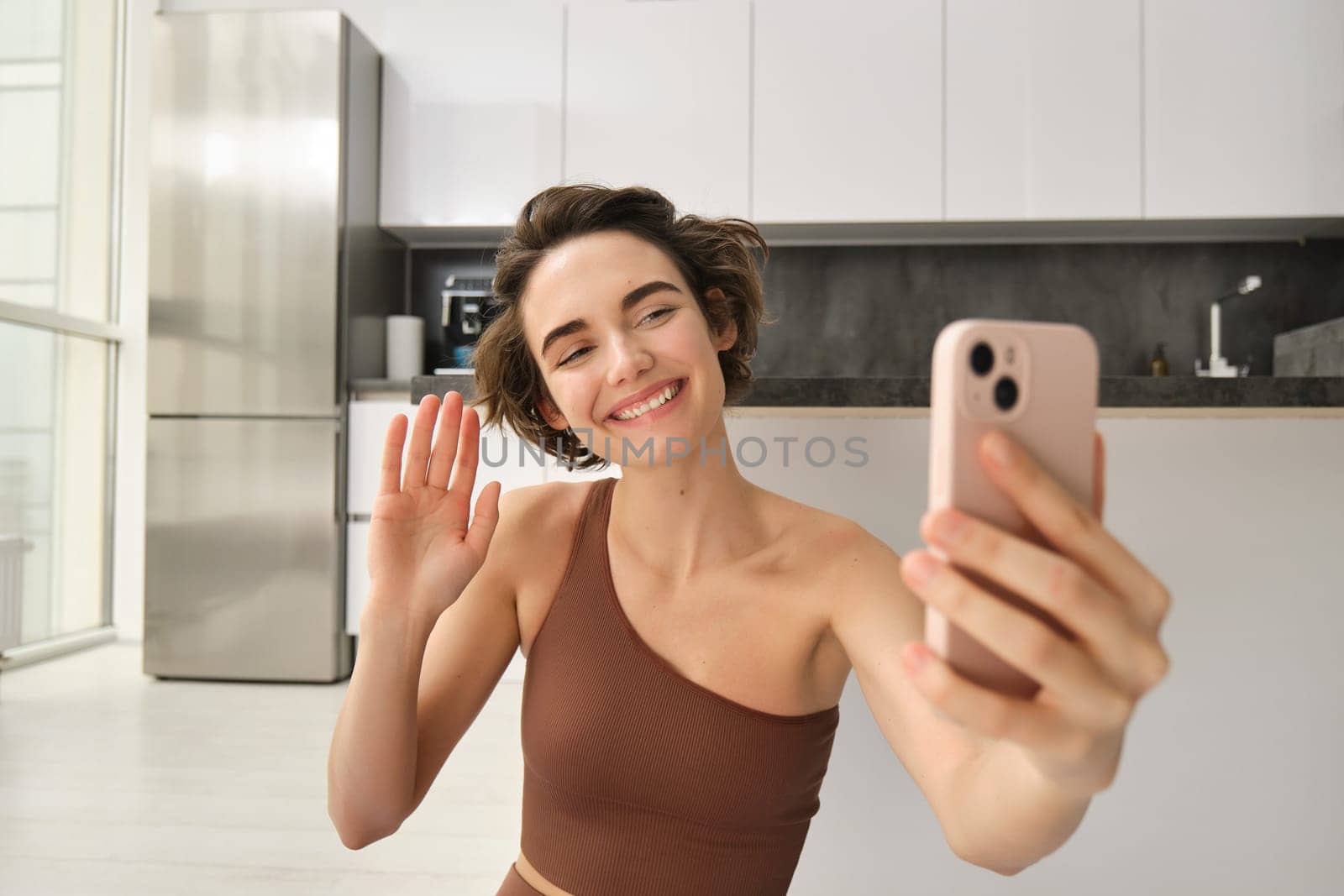 Image of young fitness girl blogger, records her workout at home, chats online while doing yoga in bright room. Woman with smartphone, joining remote yoga training via mobile phone by Benzoix