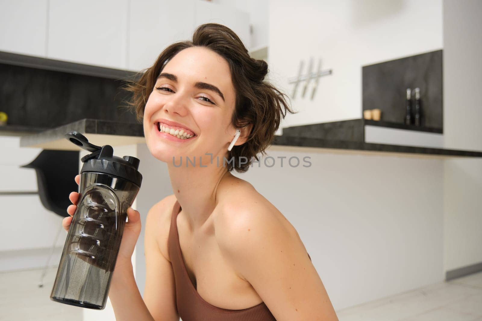 Smiling active young woman in sportswear, sits on yoga mat with water bottle, drinks stays hydrated during workout training session from home, fitness in the house on floor.