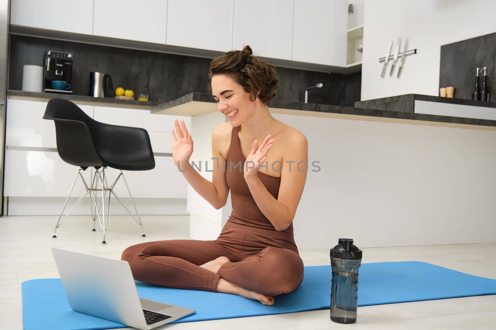 Young smiling woman sits on sports rubber mat in activewear, says hello at video chat online yoga class, using laptop during home workout training session. Copy space