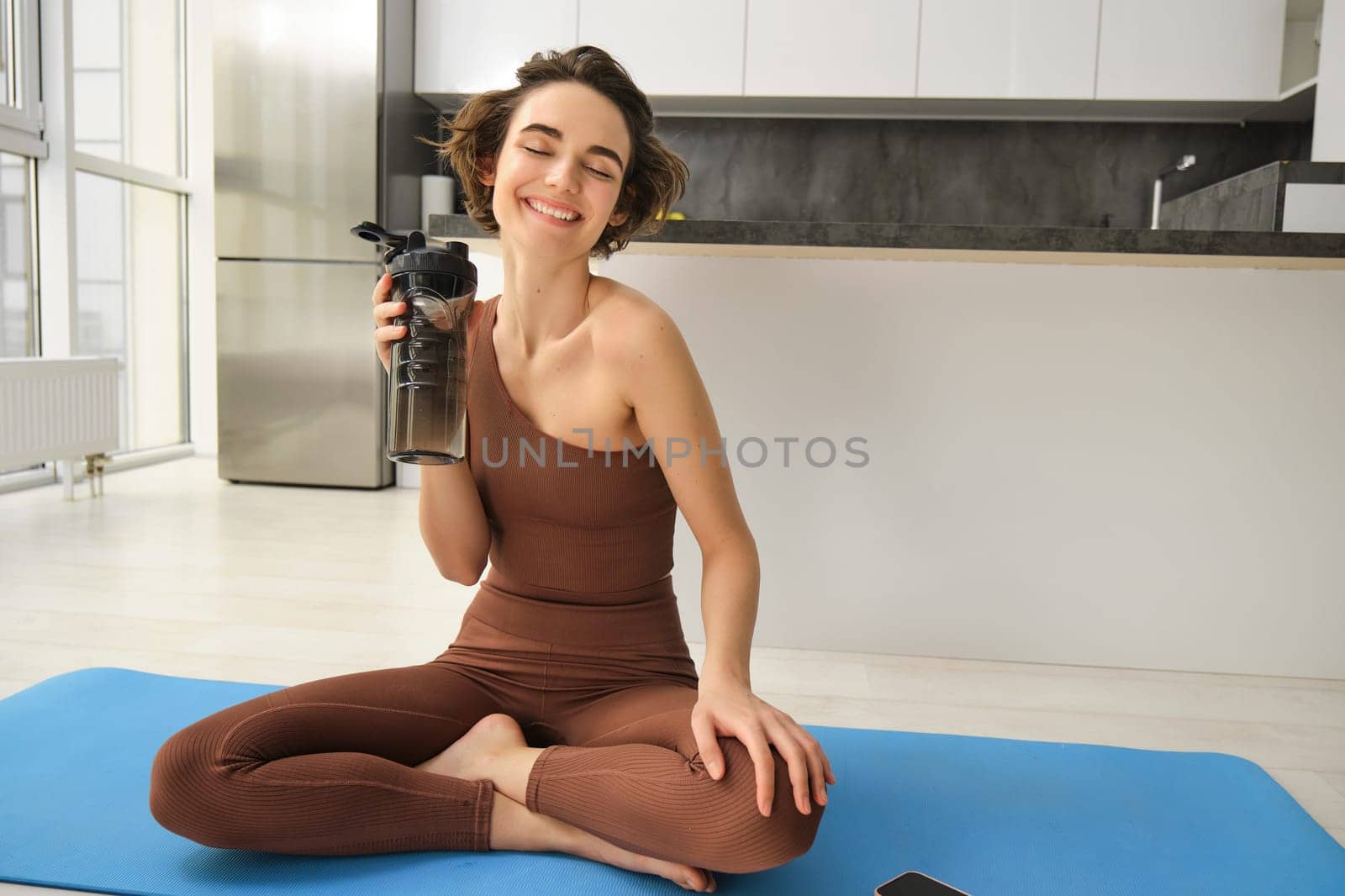 Young strong sporty athletic fitness trainer, instructor woman wearing brown tracksuit sitting on yoga mat drink water training do exercises at home gym indoor. Workout sport motivation concept.