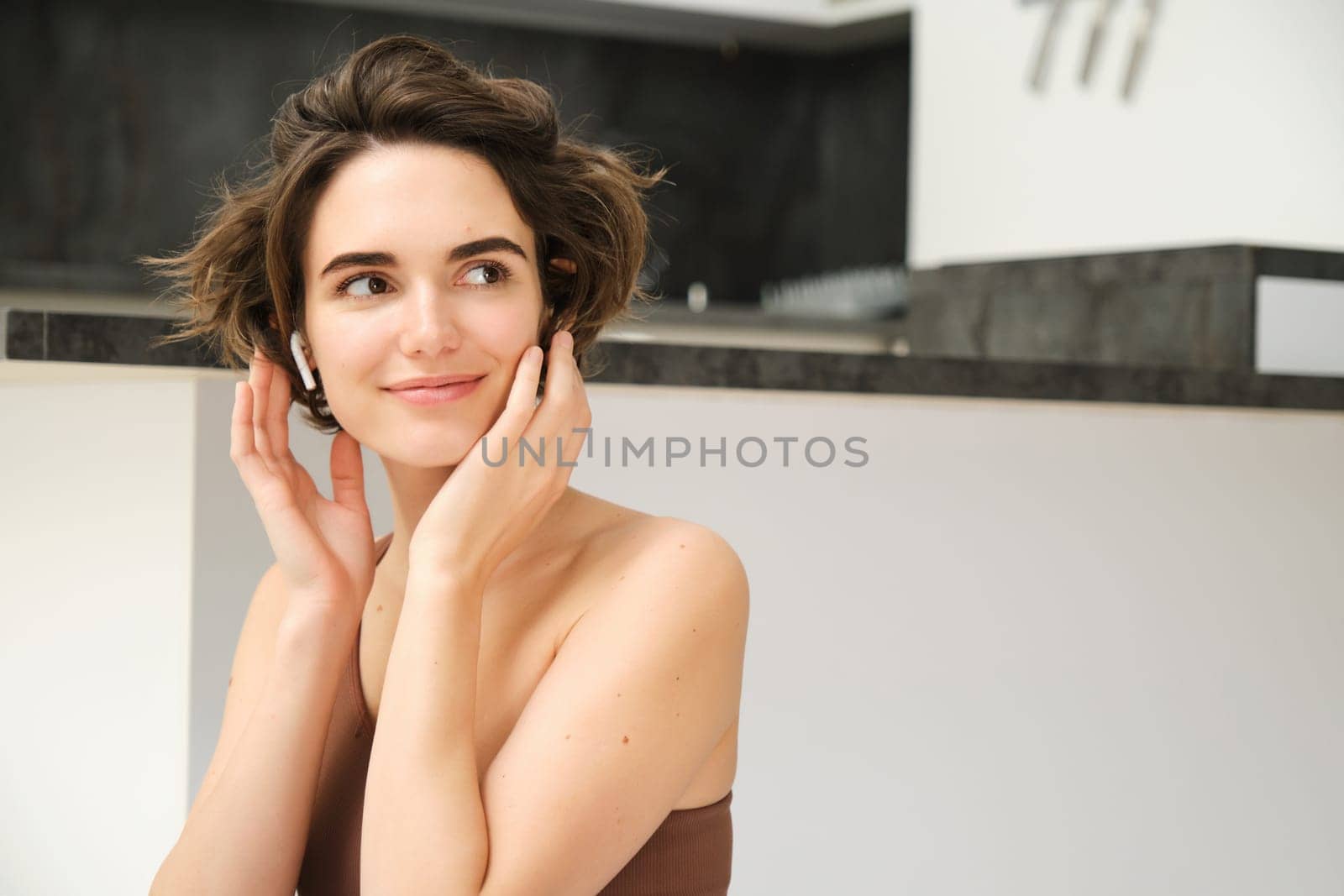 Close up of young smiling brunette woman, fitness girl in kitchen, looks aside, touches her face, listens to mindful app training session in wireless earphones, workout at home by Benzoix