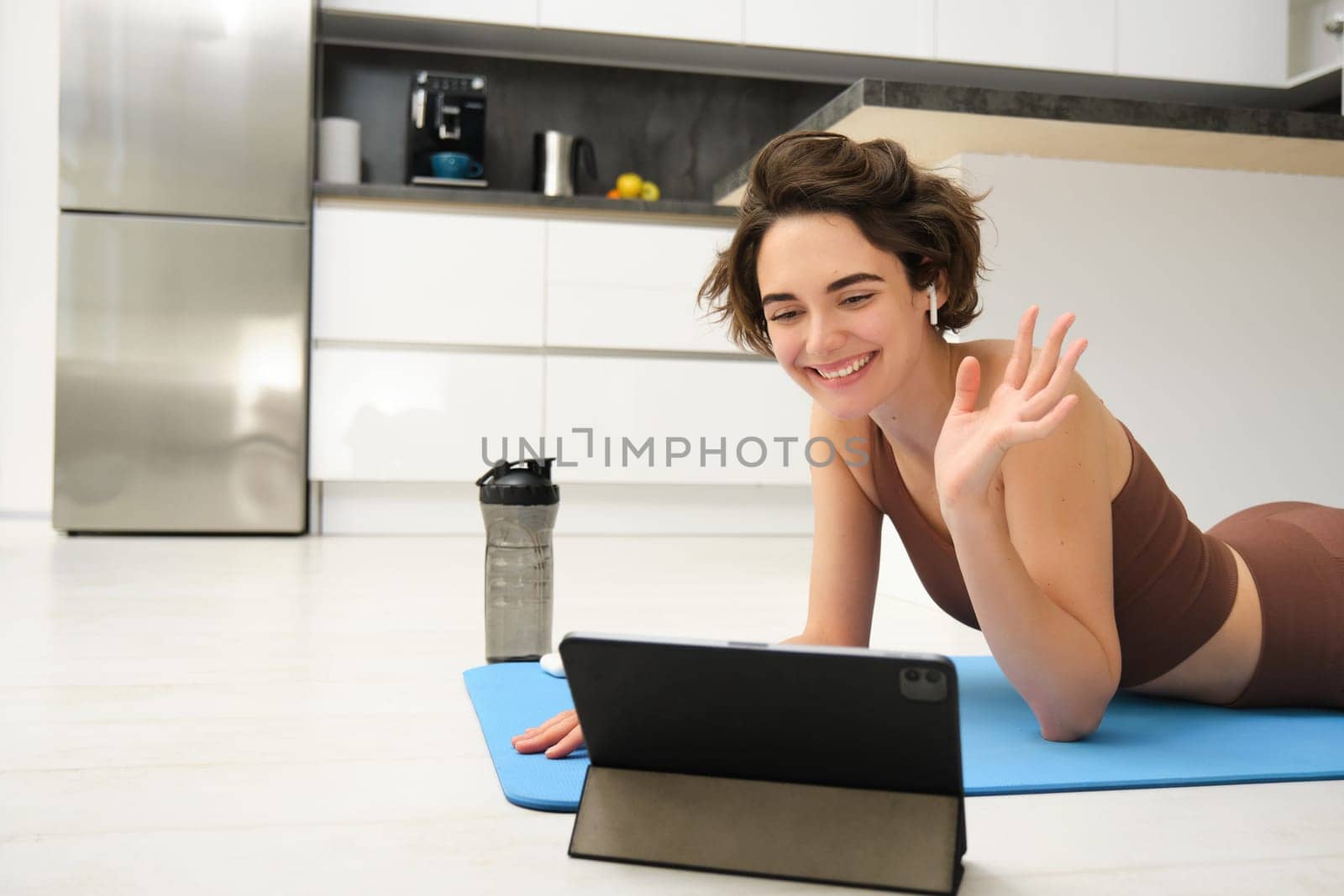 Smiling brunette woman in sportswear, waves hand, says hi at tablet, records video sports blog, fitness training session online, instructor shows workout elements, uses rubber mat at home in kitchen by Benzoix