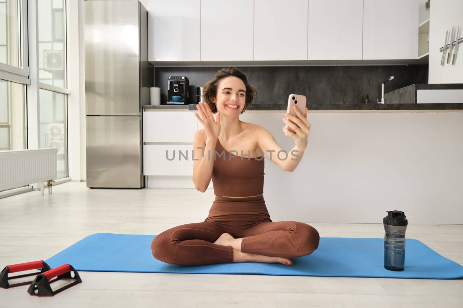 Image of young fitness girl blogger, records her workout at home, chats online while doing yoga in bright room. Woman with smartphone, joining remote yoga training via mobile phone by Benzoix