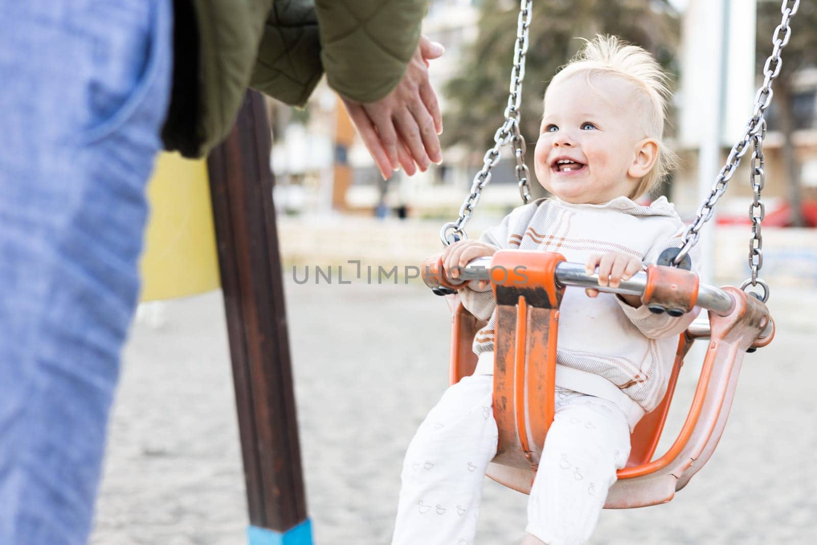 Mother pushing her cheerful infant baby boy child on a swing on sandy beach playground outdoors on nice sunny cold winter day in Malaga, Spain. by kasto