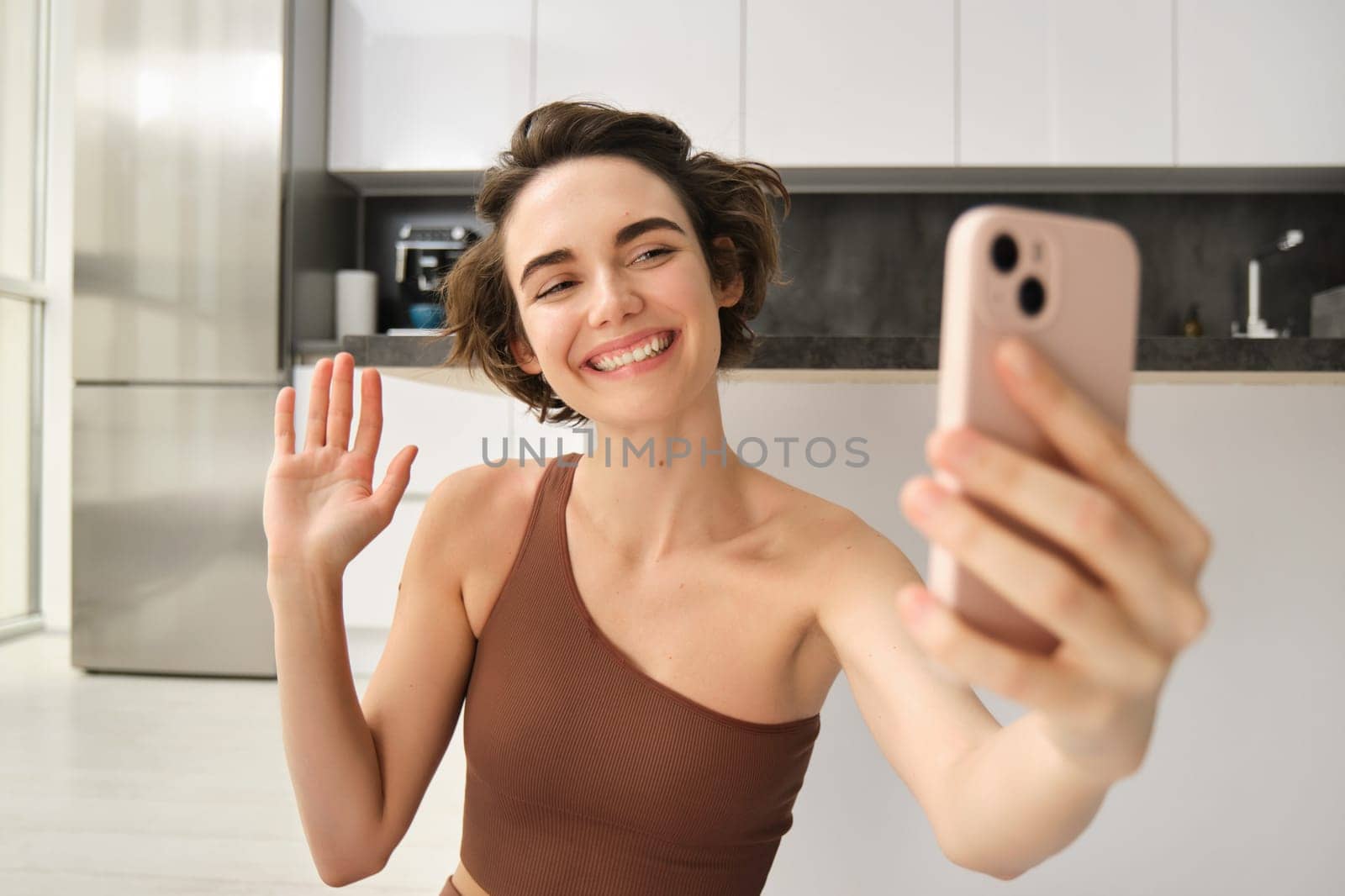 Image of young fitness girl blogger, records her workout at home, chats online while doing yoga in bright room. Woman with smartphone, joining remote yoga training via mobile phone.