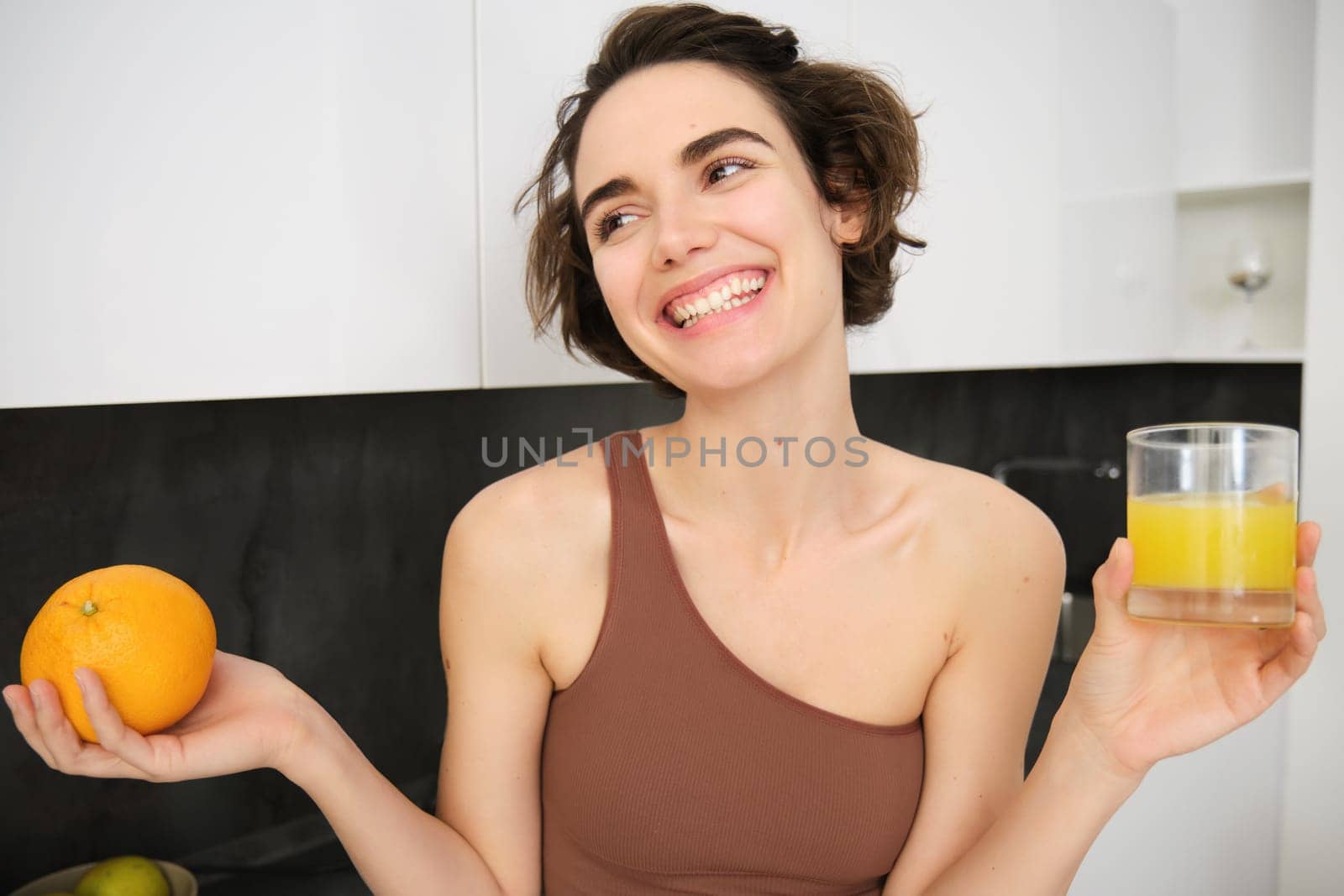 Healthy lifestyle and sport. Beautiful smiling woman, drinking fresh orange juice and holding fruit in her hand, enjoying her vitamin drink after workout at home, standing in kitchen.