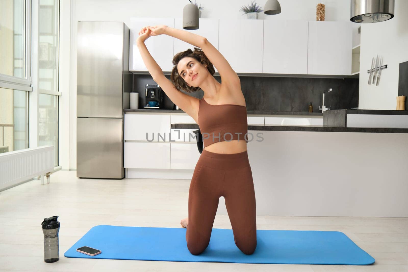 Home training. Young woman workout at home, stretching her body, doing fitness exercises on yoga rubber mat, listening to gym app instructions on smartphone by Benzoix