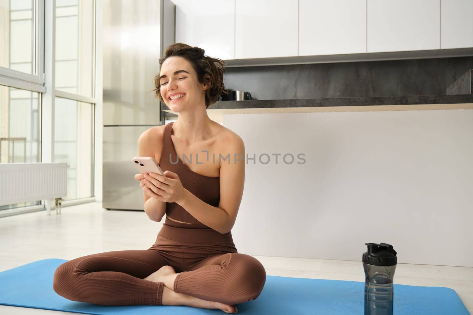Sport and women lifestyle. Young woman does workout at home, sits on yoga mat with smartphone in bright kitchen, wears activewear, uses mobile phone app for indoor exercises.
