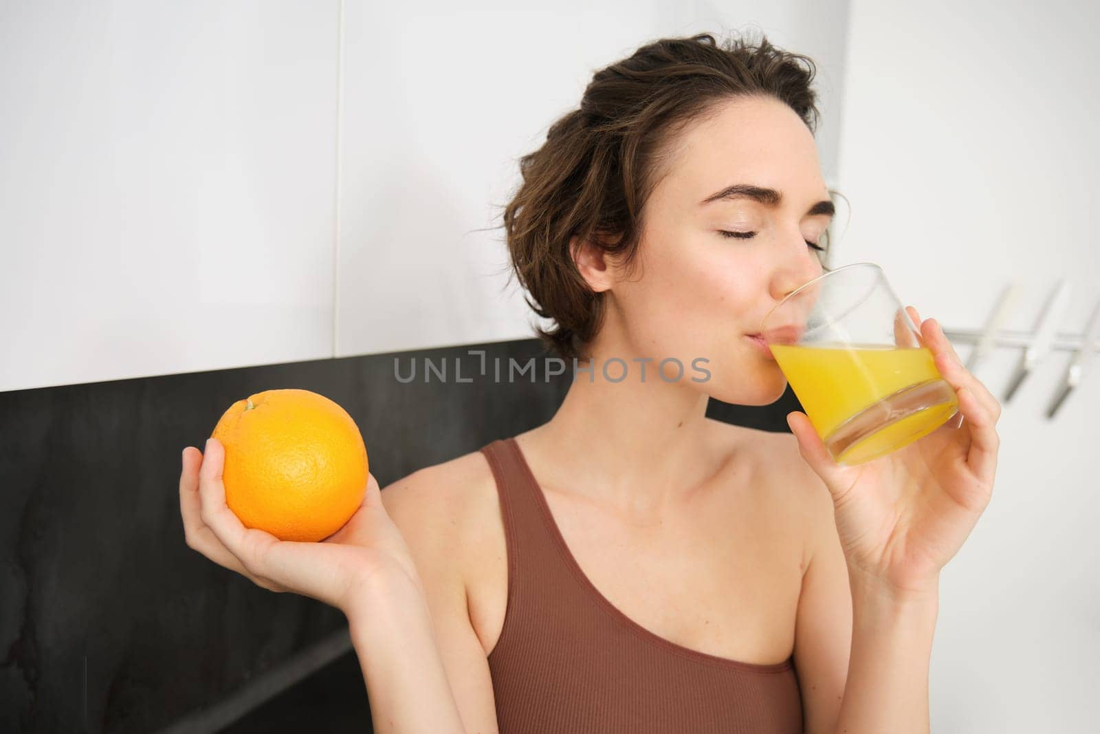 Healthy lifestyle and sport. Beautiful smiling woman, drinking fresh orange juice and holding fruit in her hand, enjoying her vitamin drink after workout at home, standing in kitchen by Benzoix