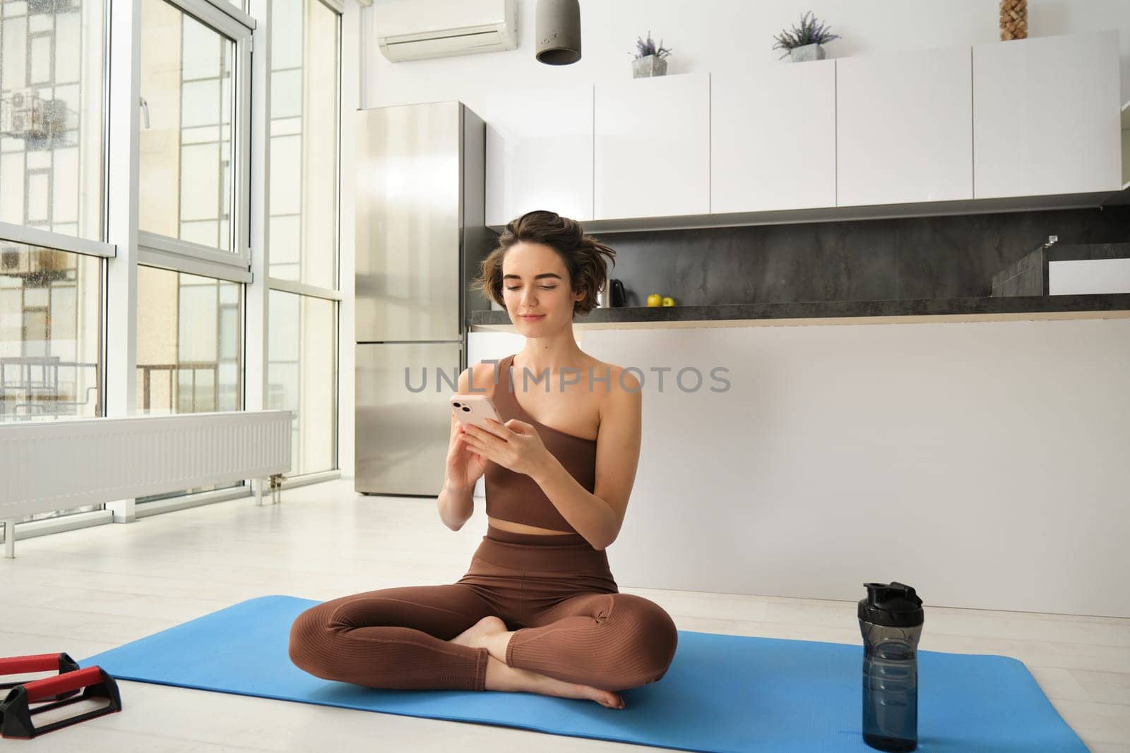 Yoga and sport at home concept. Young woman sitting in bright rome with mobile phone, doing workout on rubber mat, searching for exercises online on smartphone app, using sports equipment.