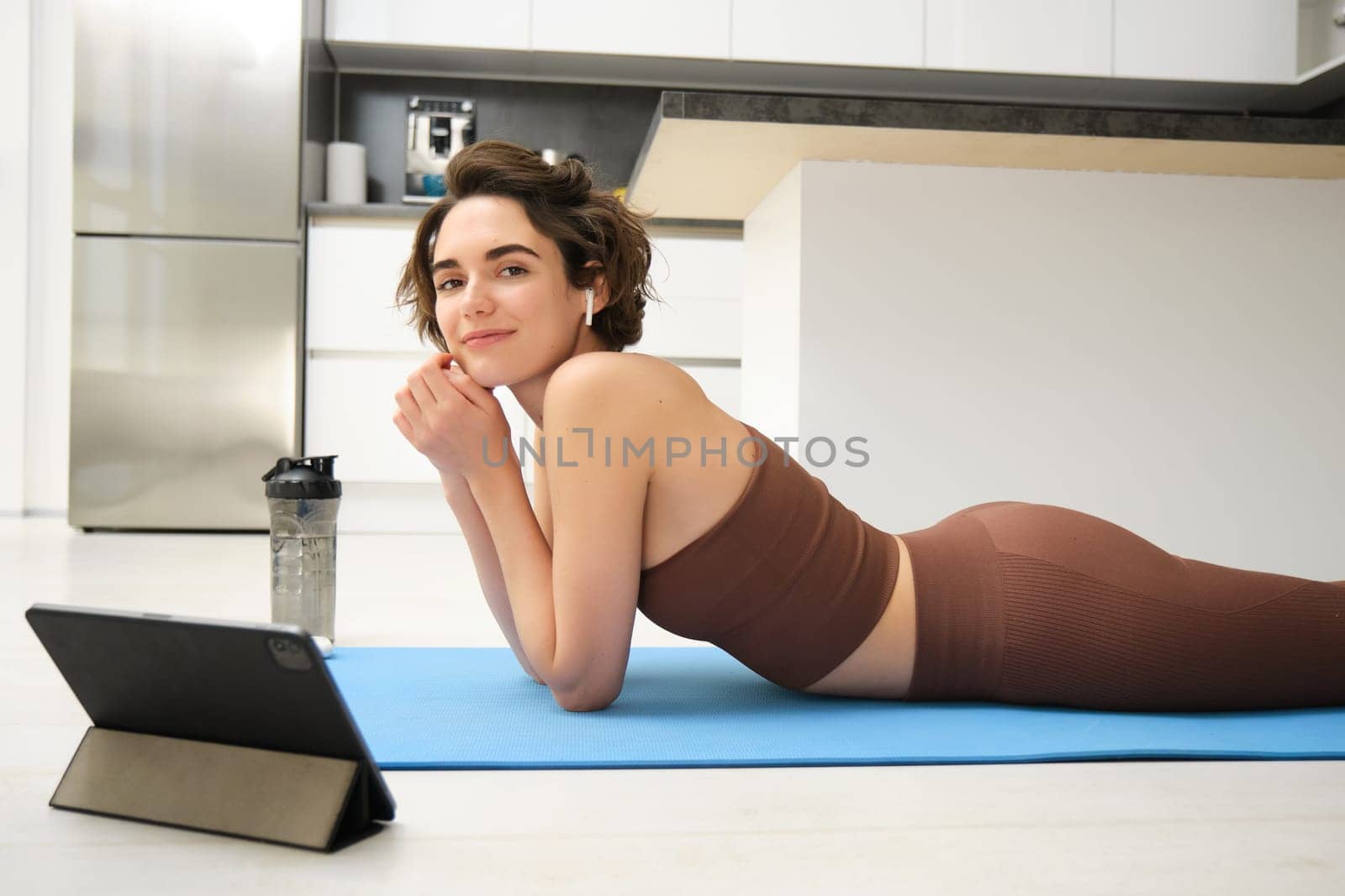 Portrait of beautiful woman in fitness sportswear, lies on floor, rubber yoga mat, uses tablet and wireless headphones, watches online sport workout tutorial, gym classes online from home.