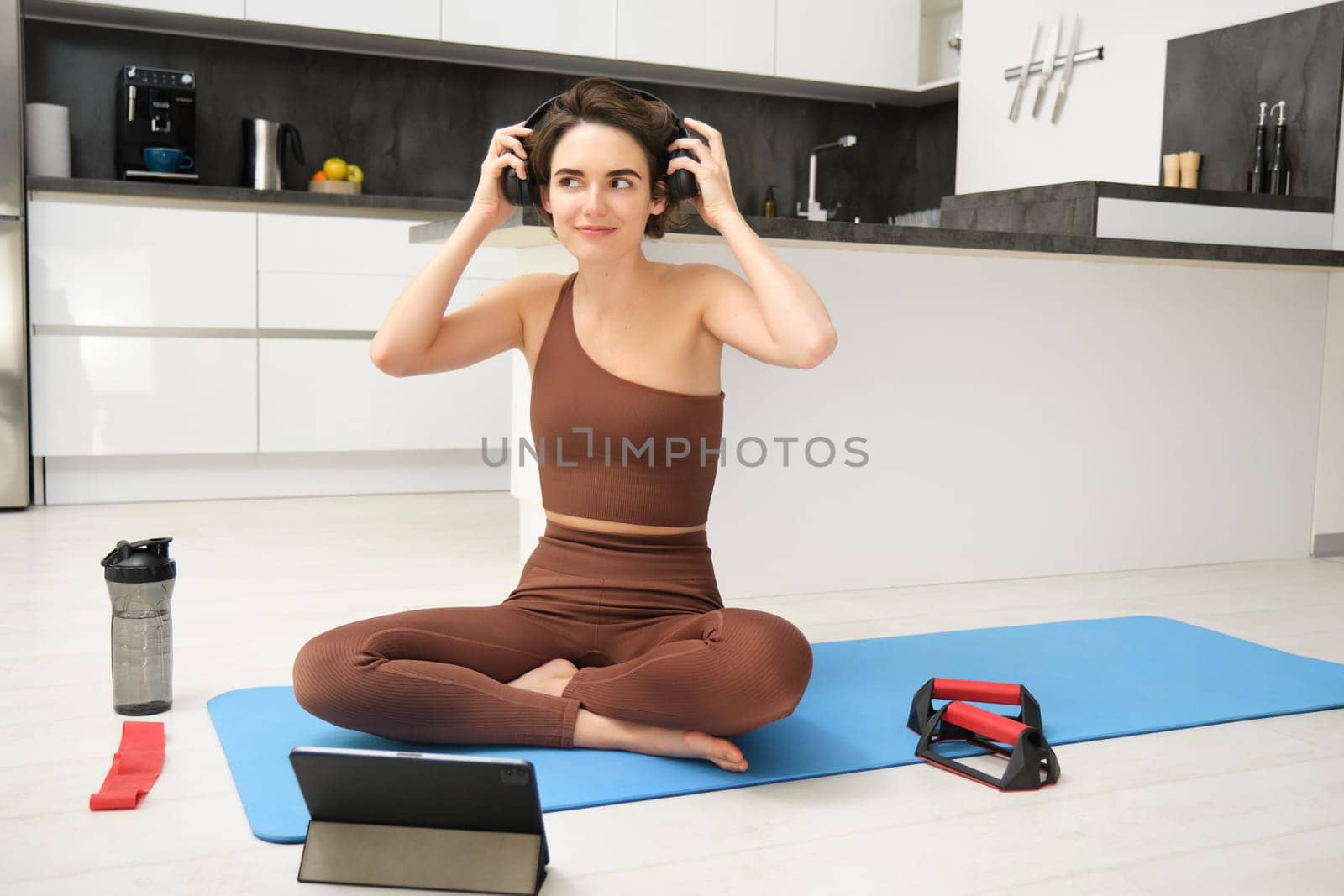 Gym at home. Young smiling woman puts on headphones in her kitchen, wearing sportswear, ready for workout, follows fitness instructor app online video on tablet, sits with exercise equipment by Benzoix