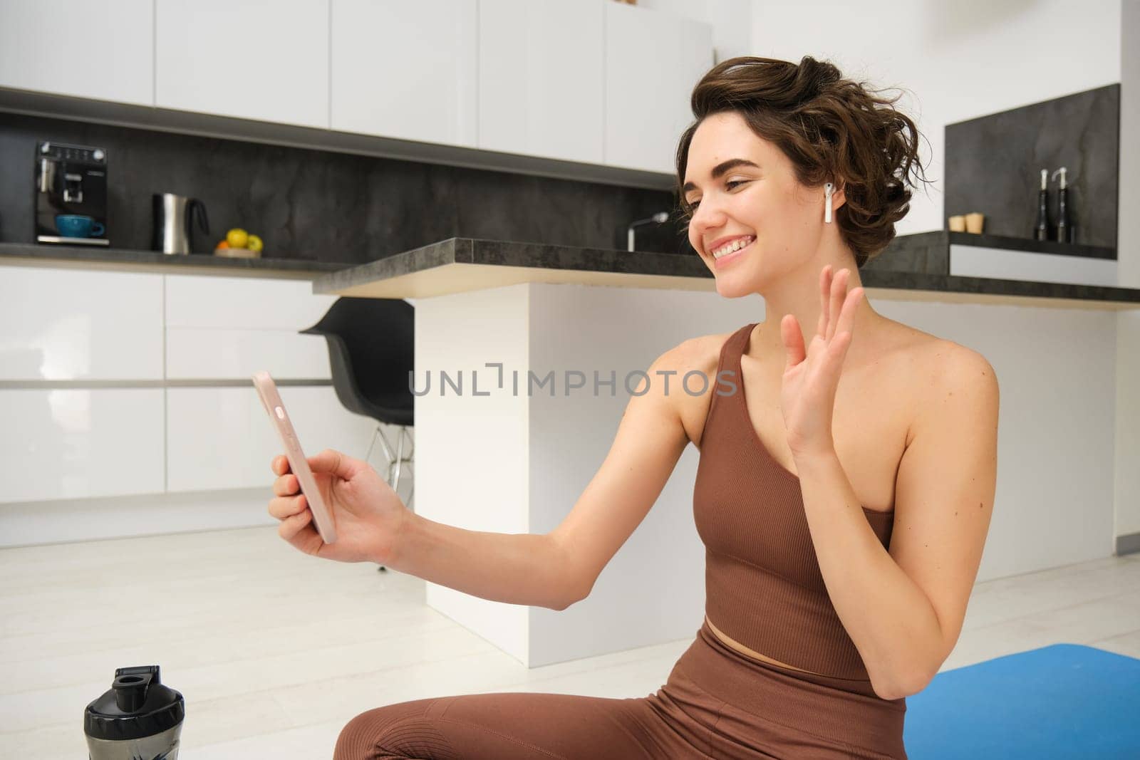 Young fitness girl at home, sport instructor in tracksuit waves hand at smartphone, sits on workout yoga mat, waves hand at mobile camera, says hi to online gym class, exercise remotely.