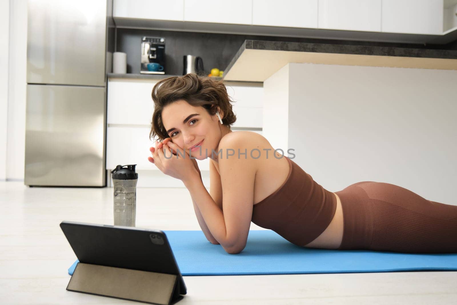 Young fitness instructor, woman does yoga, looks at tablet, watches online video pilates class, meditation training, listens in wireless headphones, lying on rubber mat, gym indoors at home by Benzoix