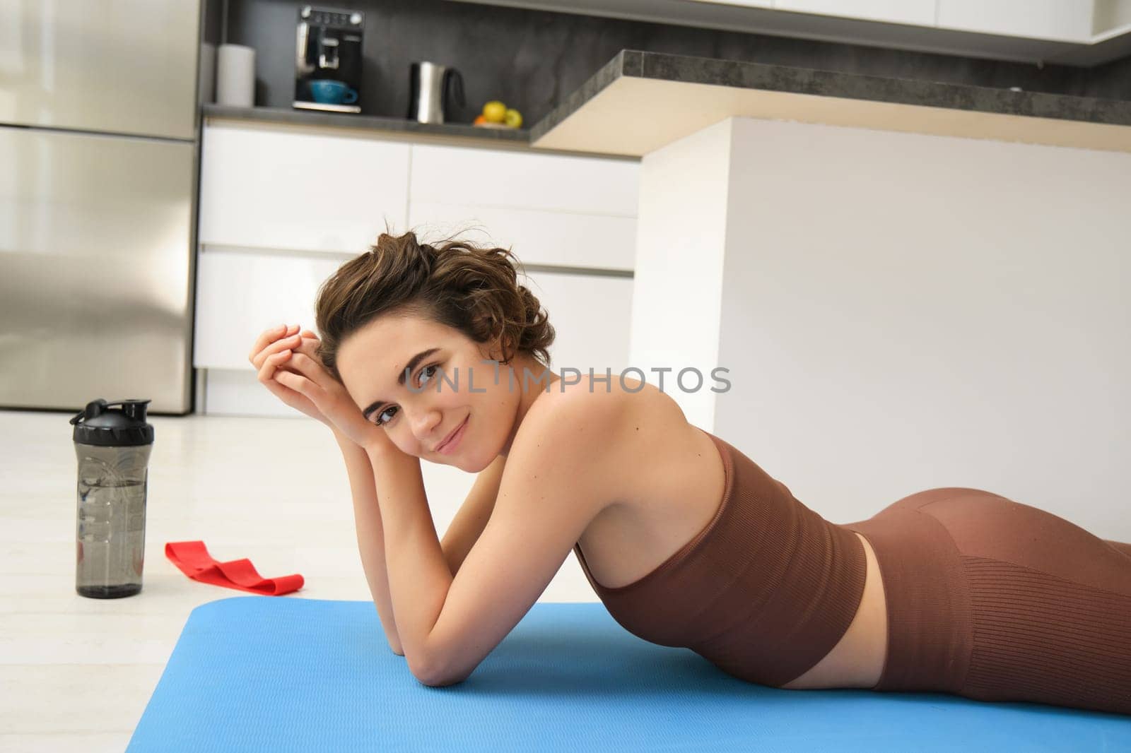 Portrait of fitness girl lying on rubber mat, warm-up before workout, doing pilates exercises at home, drinking water and doing sports, smiling at camera by Benzoix
