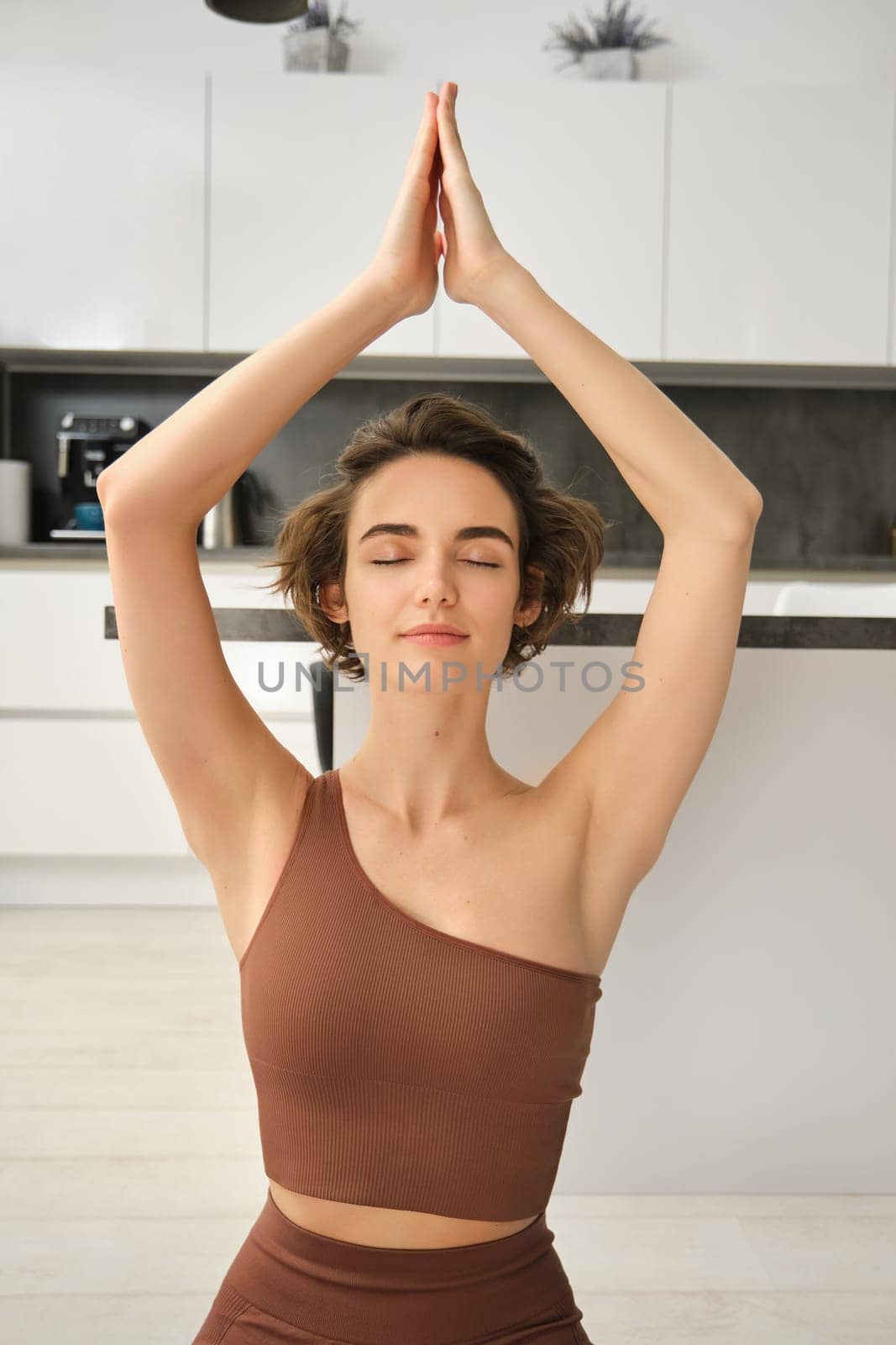 Vertical shot of young woman meditating, doing yoga at home, raising hands up and sitting in lotus pose, relaxing indoors, workout in bright room behind kitchen.