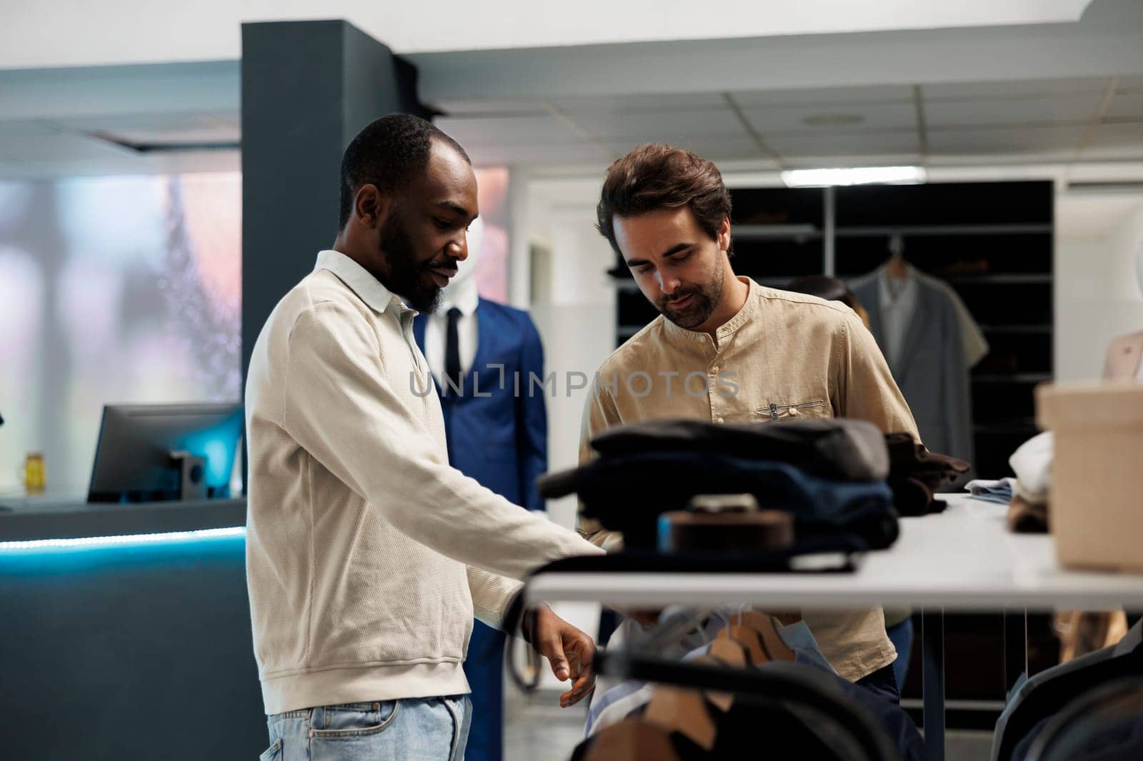 Clothing store shopper and assistant having conversation about fashion trends while exploring apparel rack. African american man asking boutique worker for help in choosing outfit