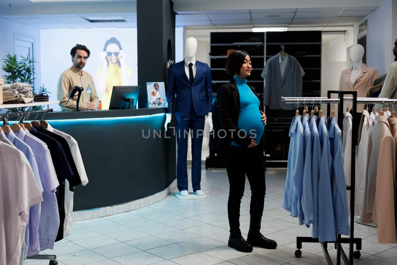 Young pregnant asian woman searching for maternity clothes in mall fashion boutique. Expectant mom standing near rack with hanging apparel in shopping center, choosing garment