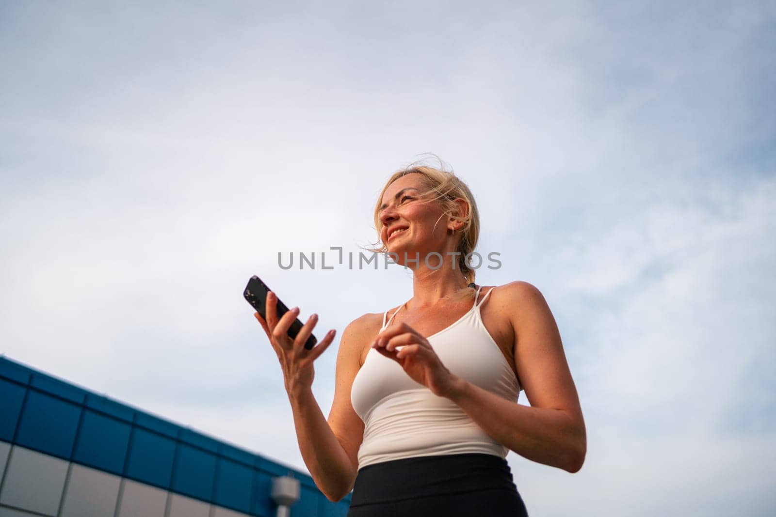 Beautiful blonde caucasian woman using phone while doing sports outdoors. Healthy fitness lifestyle, summer workout in city. Using technology for tracking activity by troyan