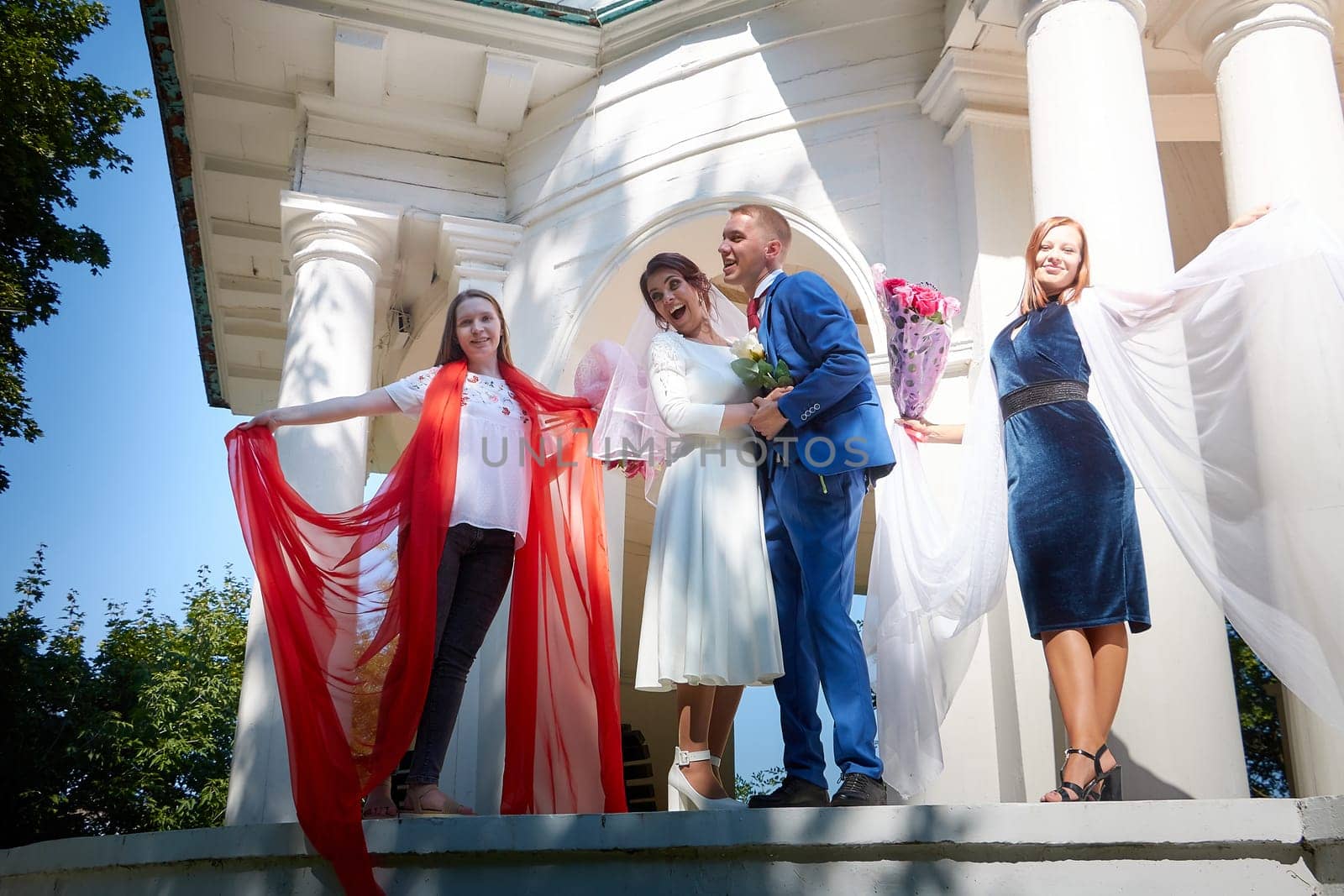 Kirov, Russia - August 10, 2021: Groom and bride walking in the park on sunny summer day and bridesmaids with beautiful fabrics accompanying them. Friends and guests at wedding with newlyweds outdoors by keleny