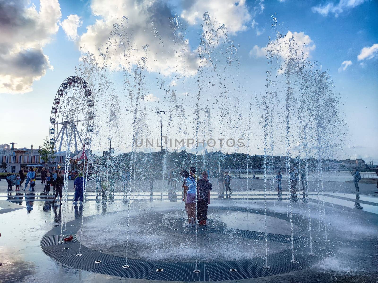 Cheboksary, Russia - June 01, 2023: Water jets of a fountain from the sidewalk and people and children cheerfully rejoice in city. Resting place for townspeople on a hot summer evening. Partial focus