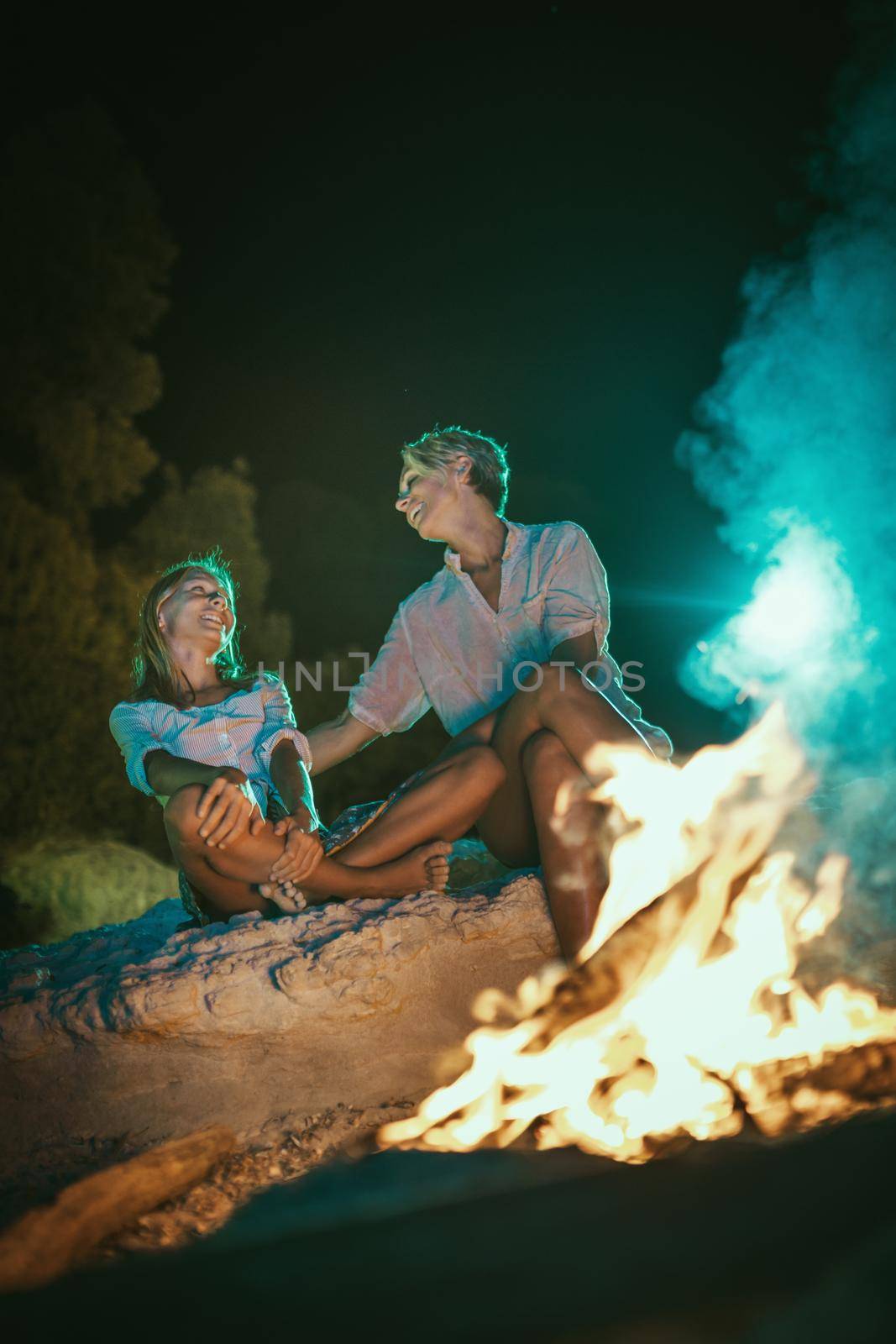 The happy smiling daughter and her mother are sitting on the sandy seashore by the fire in the evening.