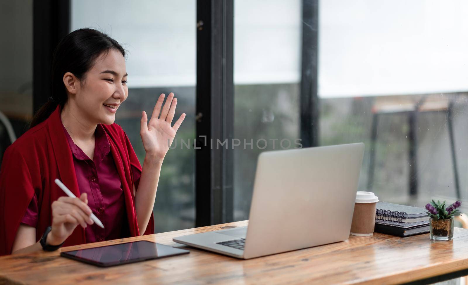 Close up photo of positive cheerful asian girl have online video call broadcast say hi wave hand sit table use laptop in house.