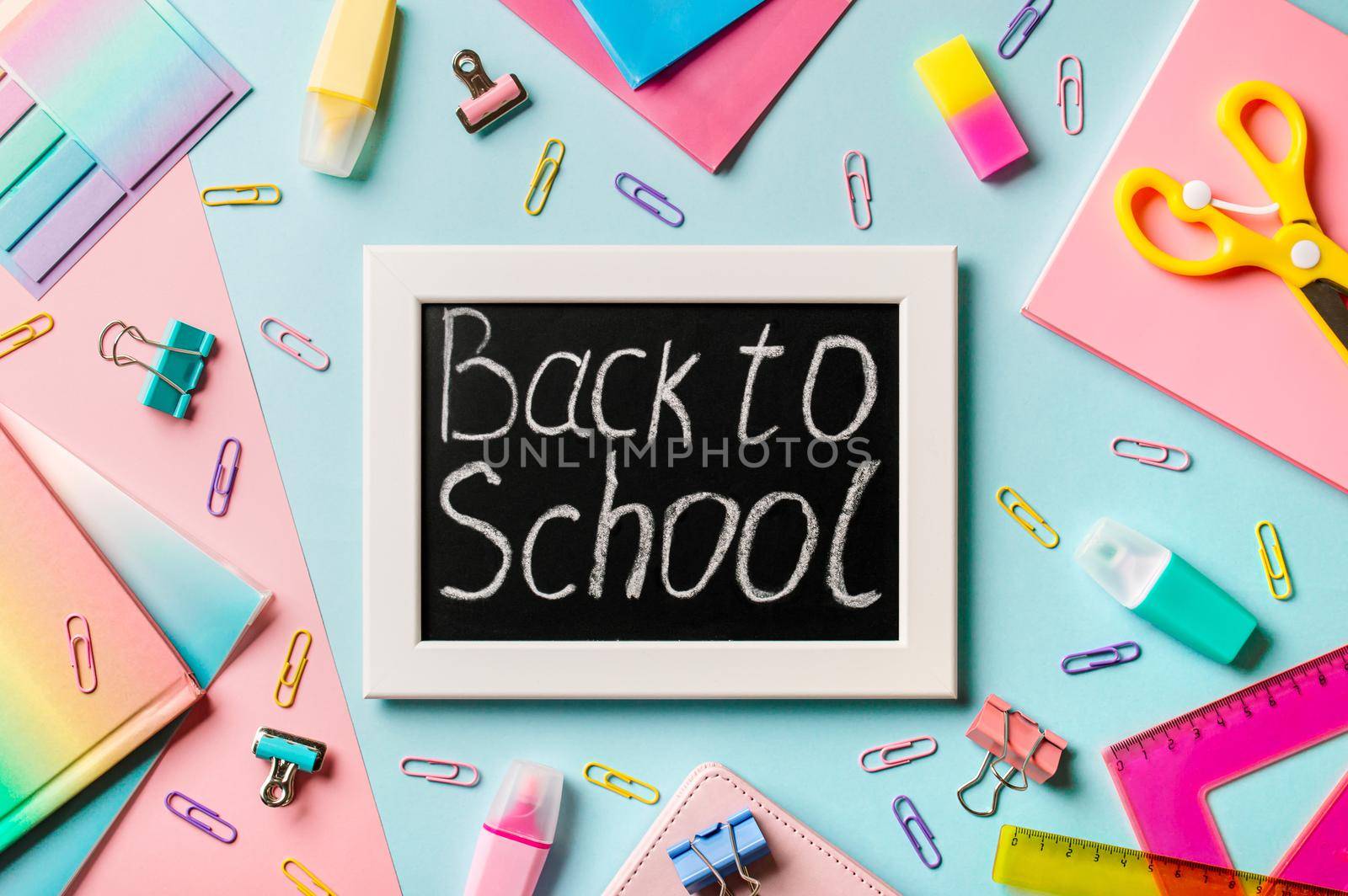 Blackboard with letters Back to school made with chalk. School supplies, pencils, scissors and colored books on blue and pink background. Top view or flat lay. Back to school concept