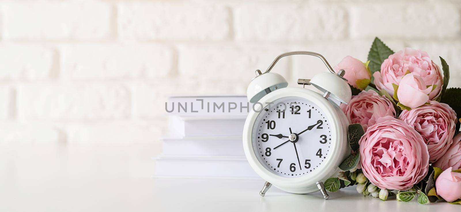 White retro alarm clock on white brick wall with books and peonies with copy space by Desperada