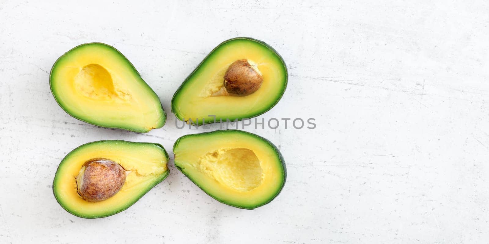 Overhead shot - four avocado halves, two with seed visible, on white working board; wide banner space for text at right side. by Ivanko