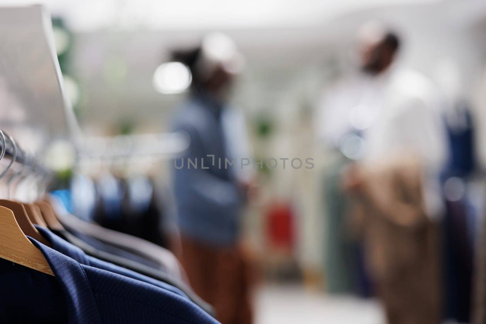 Male shirts hanging on rack in shopping mall with customer and assistant choosing clothes on blurred background. Menswear on hangers in fashion boutique close up selective focus