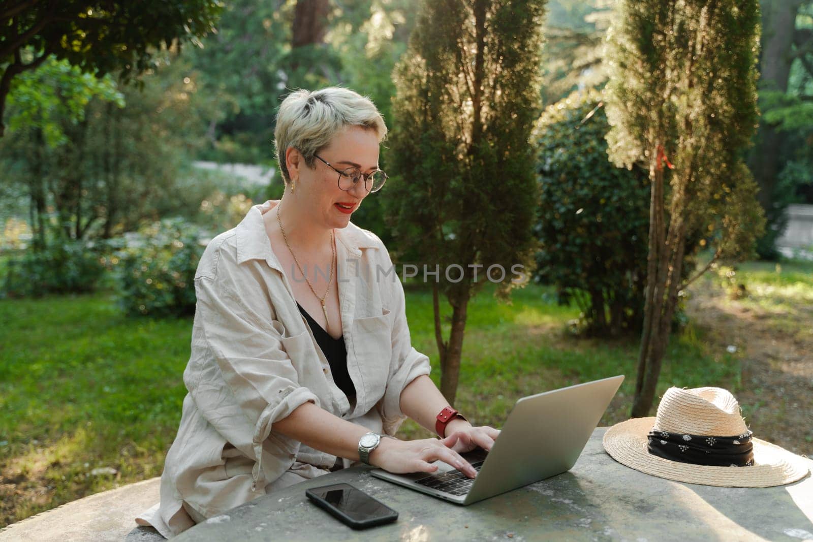 woman freelancer in glasses works at a computer at a white table in nature and spends her day productively.