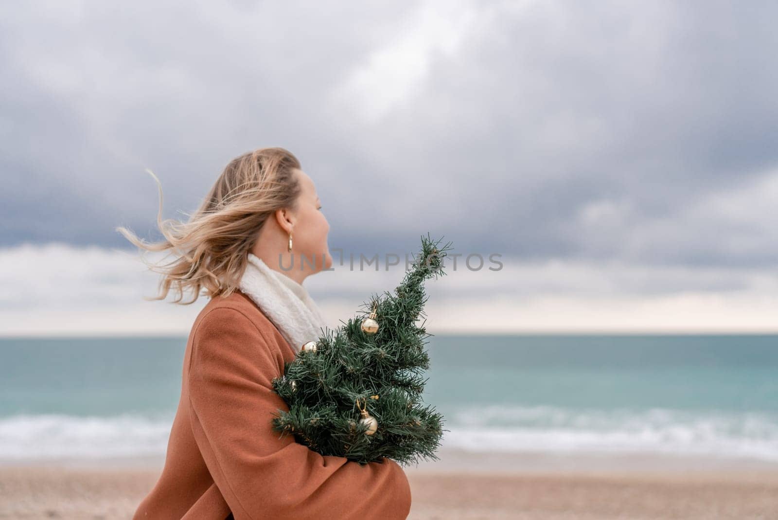 Blond woman Christmas sea. Christmas portrait of a happy woman walking along the beach and holding a Christmas tree on her shoulder. She is wearing a brown coat and a white suit