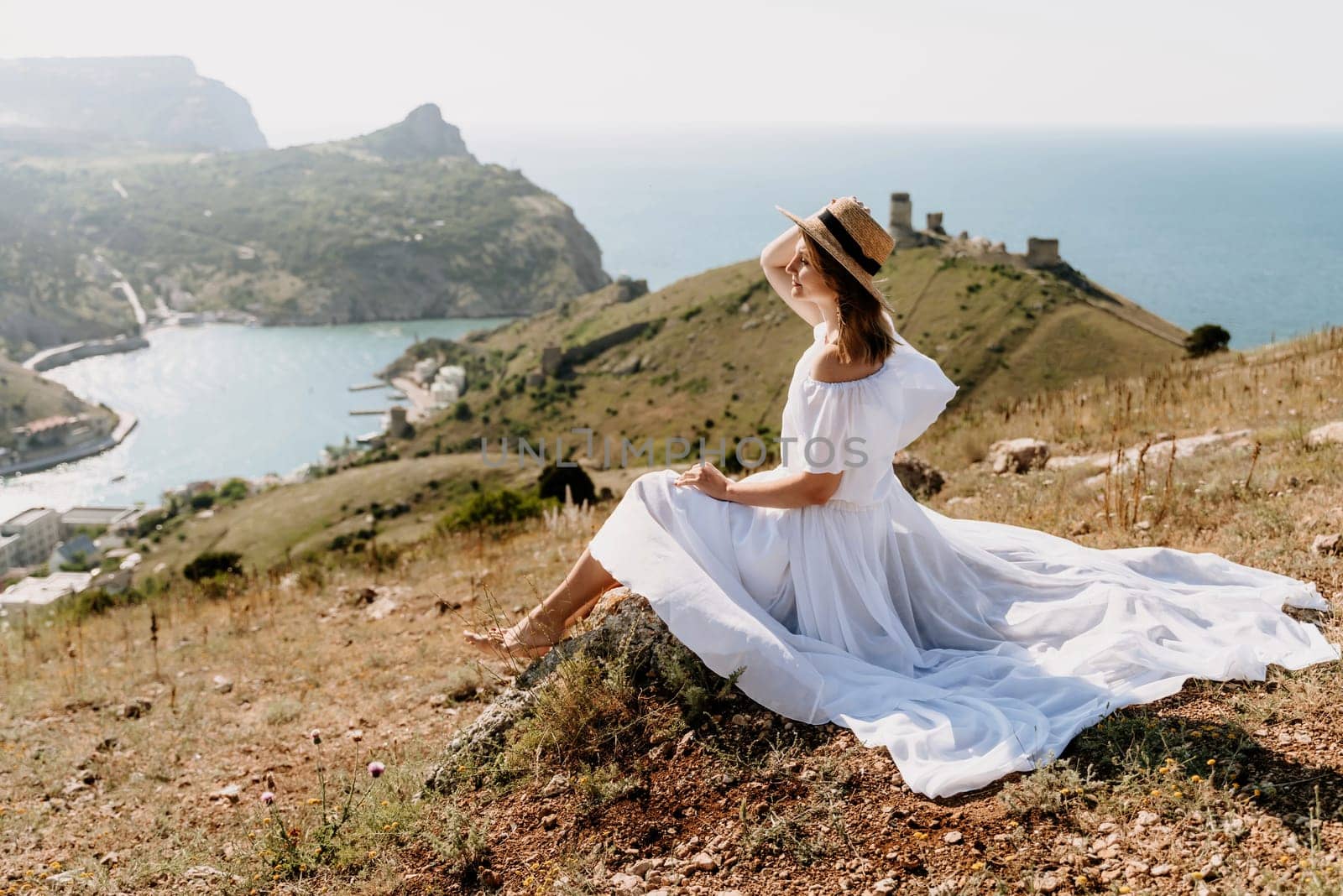 Happy woman in a white dress and hat stands on a rocky cliff above the sea, with the beautiful silhouette of hills in thick fog in the background