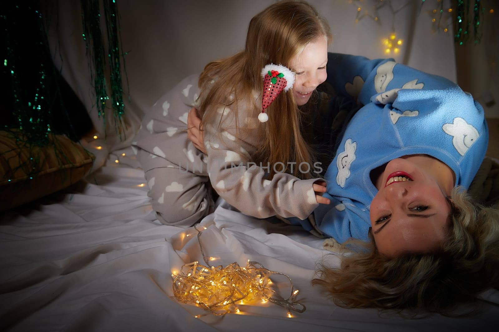 Cute mother and daughter in pajamas lie on soft blanket and having fun in room with Christmas garlands and white background. Tradition of decorating house for holiday. Happy childhood and motherhood