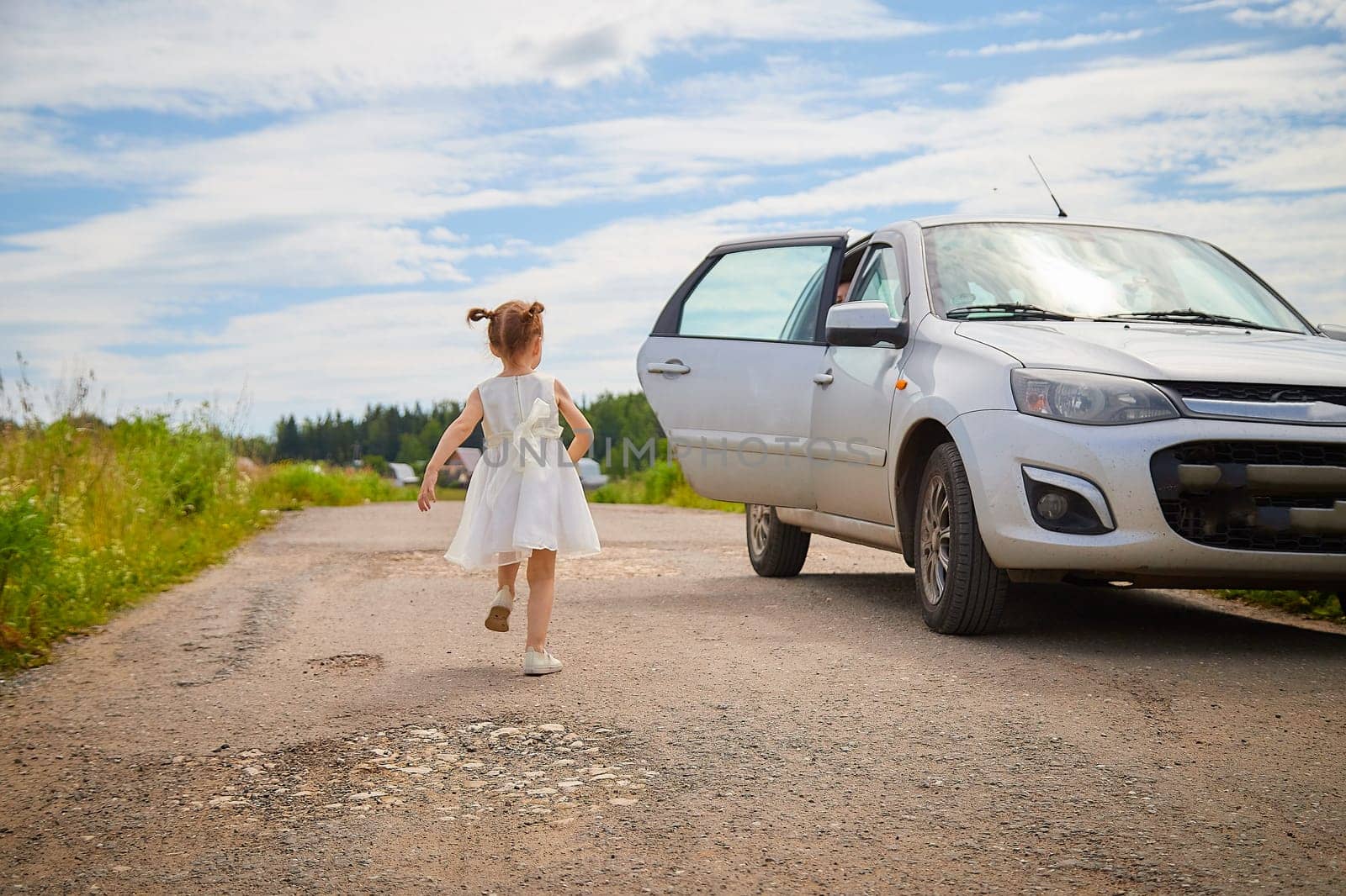 Portrait of little girl with Asian eyes on a road near car in a meadow or field with grass and flowers on sunny summer day. Rural, rustic journey