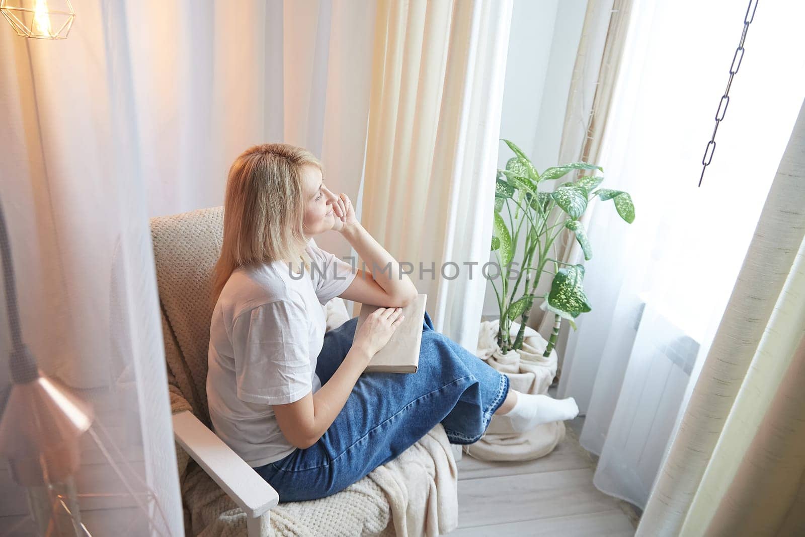 Young blonde woman with book on chair near window at home. Student girl is resting and reading textbook at home. Nice atmosphere. The concept of a cozy home by keleny