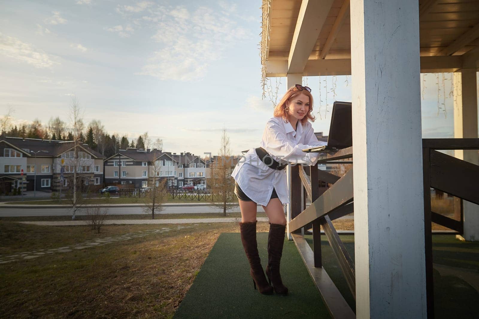 Beautiful redhead moden girl working by laptop in gazebo on autumn, summer or spring day. Businesswoman, student, freelancer or manager works outdoors in natural landscape in a village, city or town