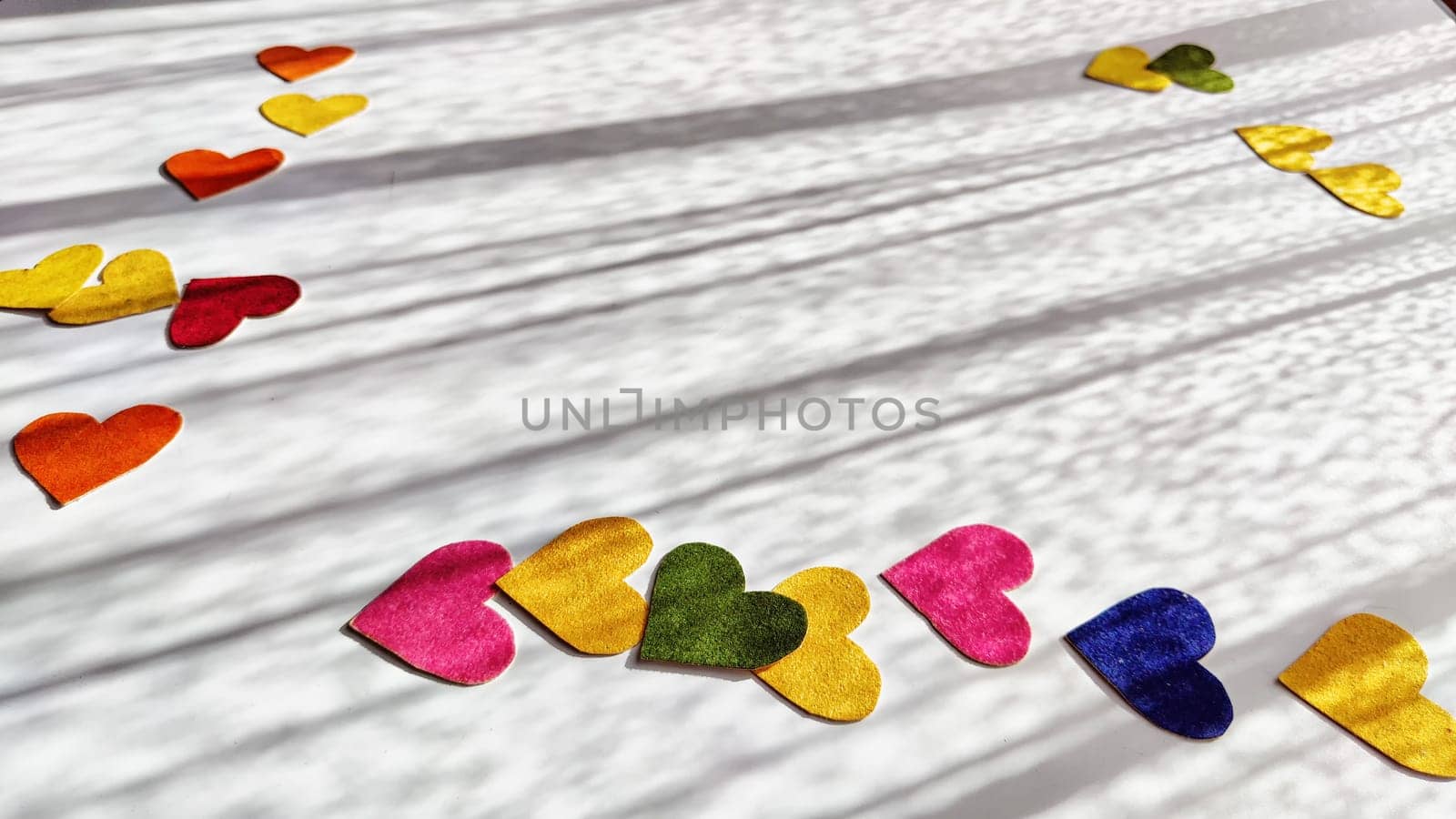 Multicolored hearts made of rough velvet paper on white surface with stripes of shadow and light from the sun. Abstract background, texture, copy space