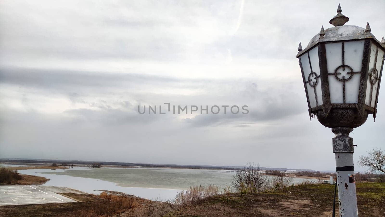 Landscape with the water of a lake, river, bay or reservoir with gray water, gloomy sky, clouds, the shore and beautiful lantern on an autumn, spring or winter day. View from a height