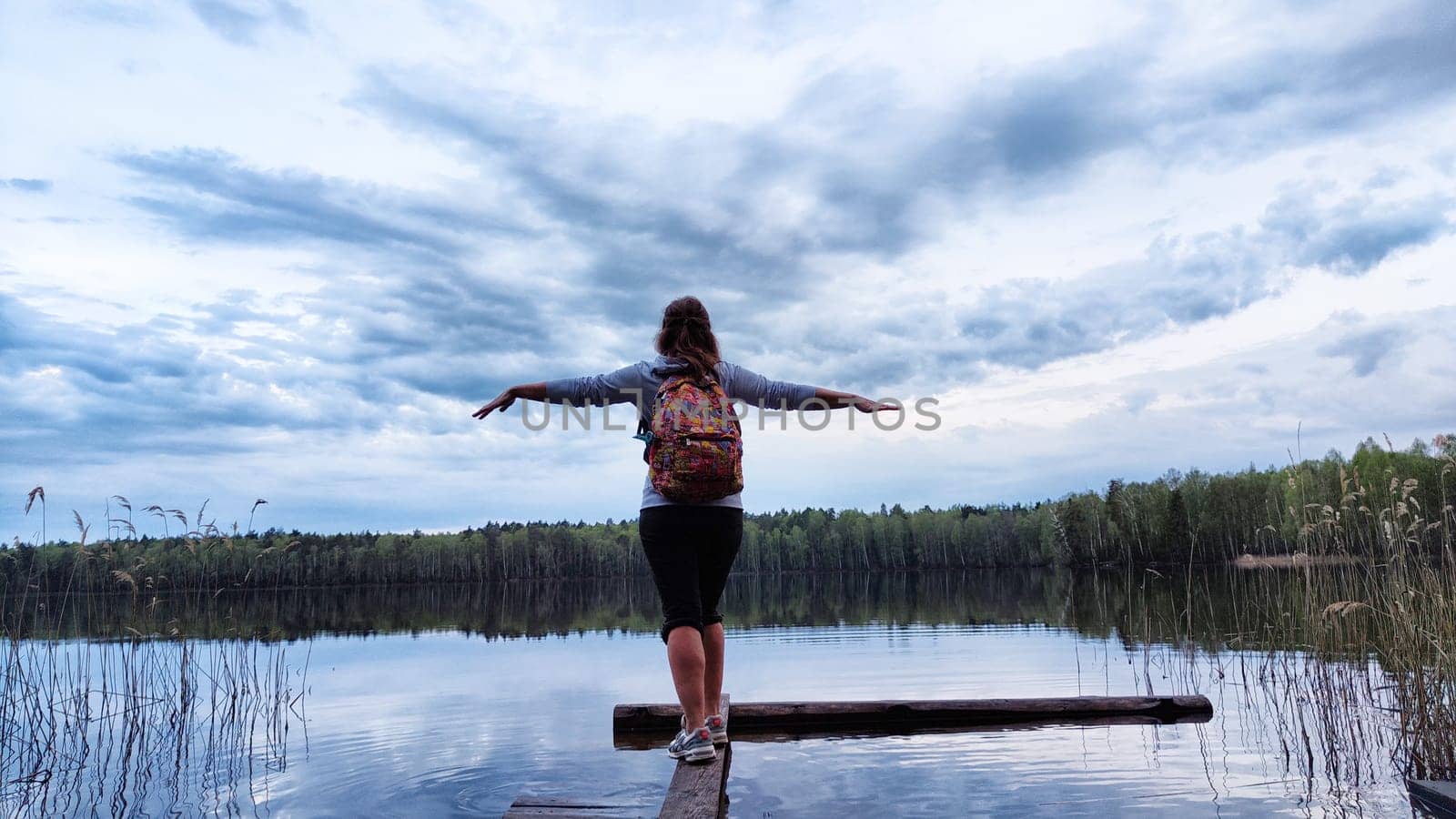 Girl or woman walks along old wooden pier, with aake in background during evening and landscape featuring water, forest and clouds in sky. Tourist in natural landscape. Concept of tourism and freedom