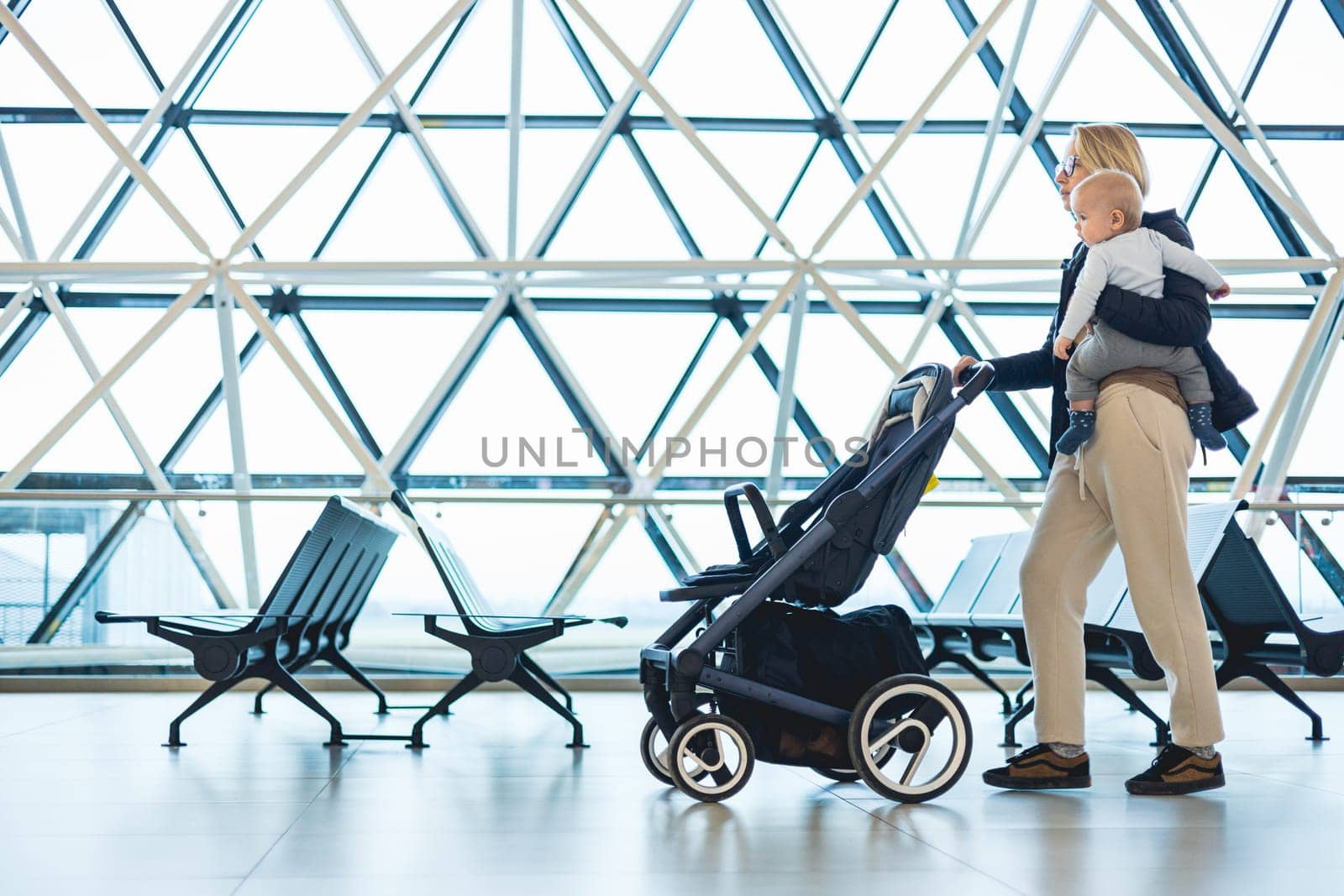 Mother carying his infant baby boy child, pushing stroller at airport departure terminal moving to boarding gates to board an airplane. Family travel with baby concept. by kasto