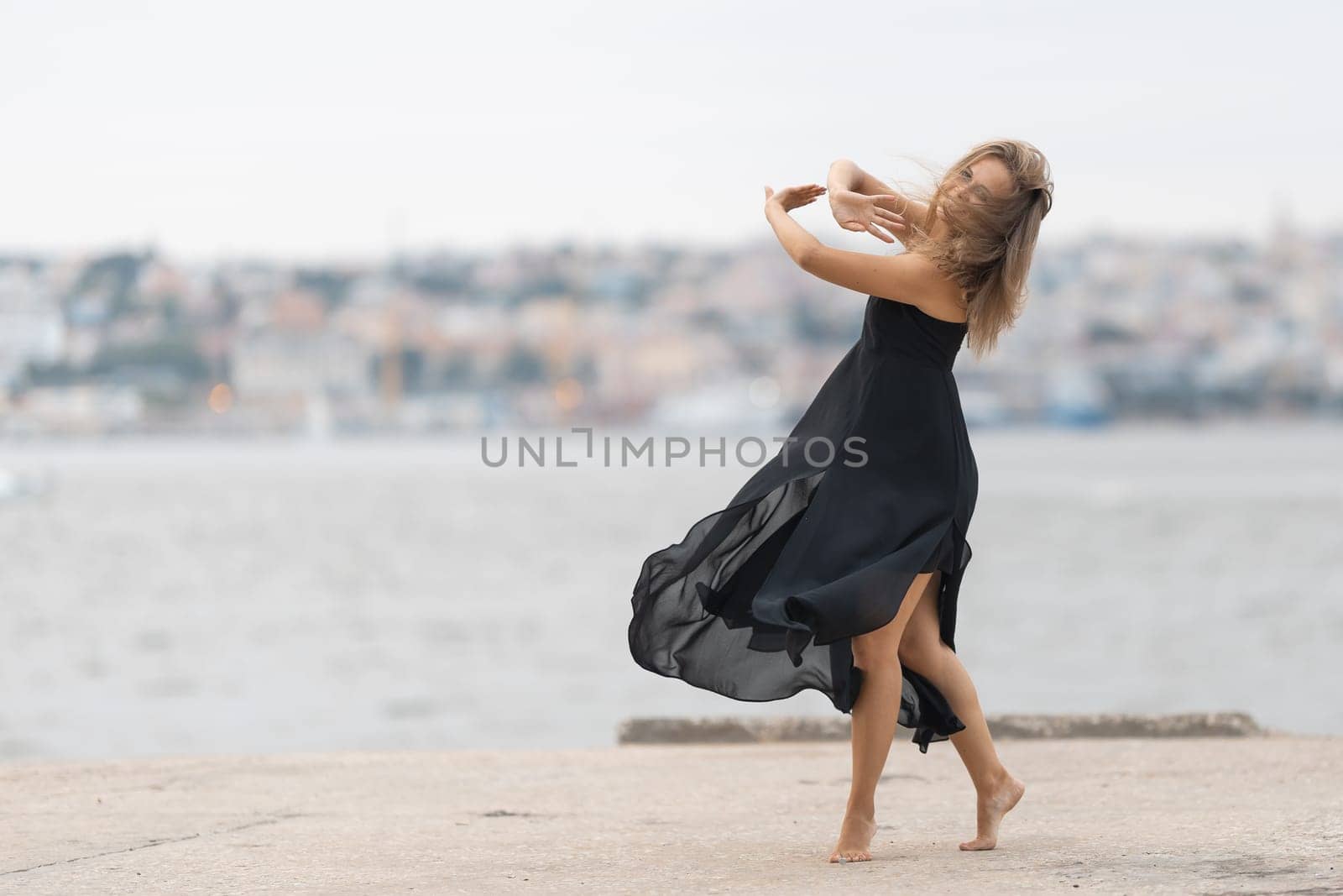 An adult woman in black dress dancing ballet on the pier in cloudy weather by Studia72